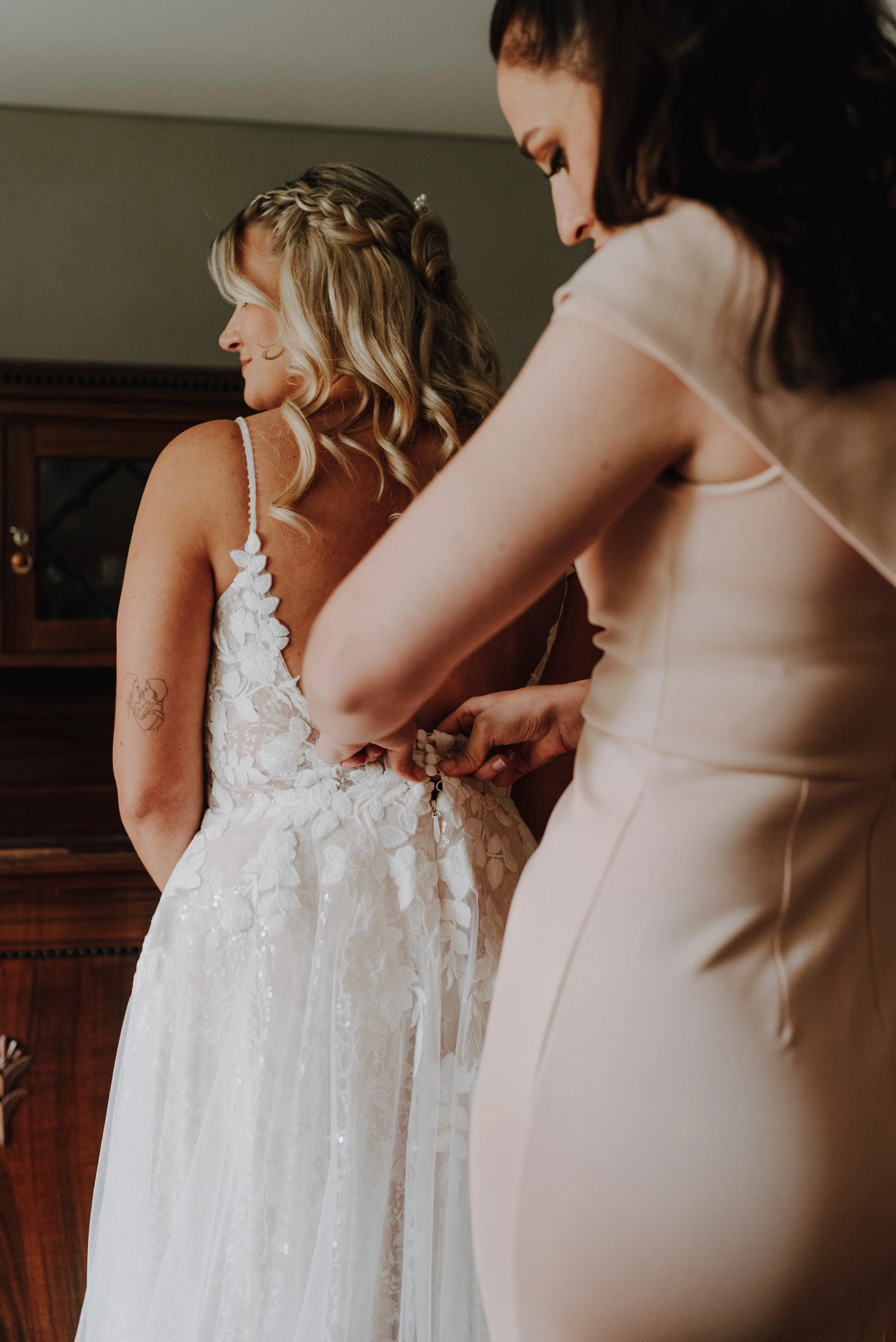 Eine Braut wird von einer Helferin für die Hochzeit vorbereitet, während sie den Knopf an ihrem Hochzeitskleid schließt.