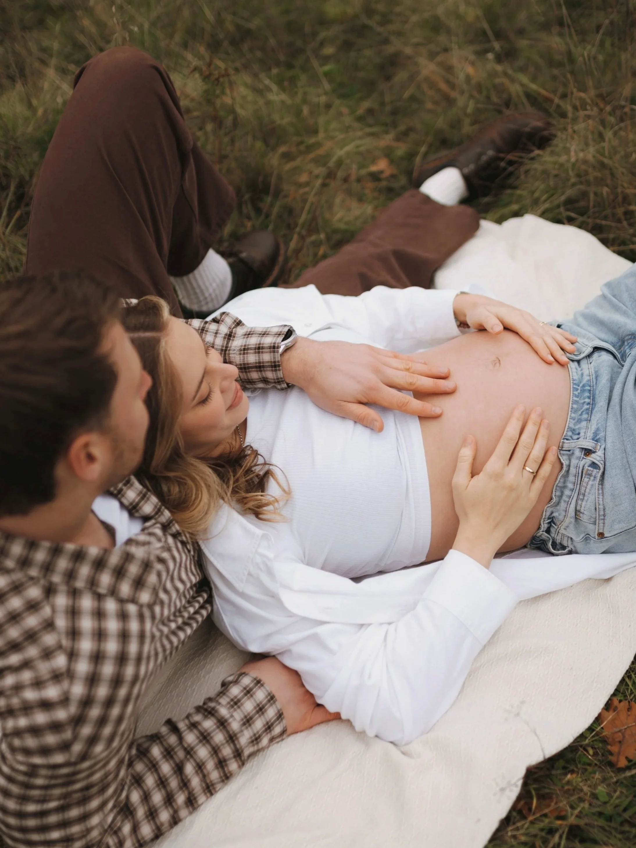 Paar liegt auf einer Decke im Freien, die Frau berührt den Babybauch, während der Mann neben ihr sitzt, umgeben von Natur.