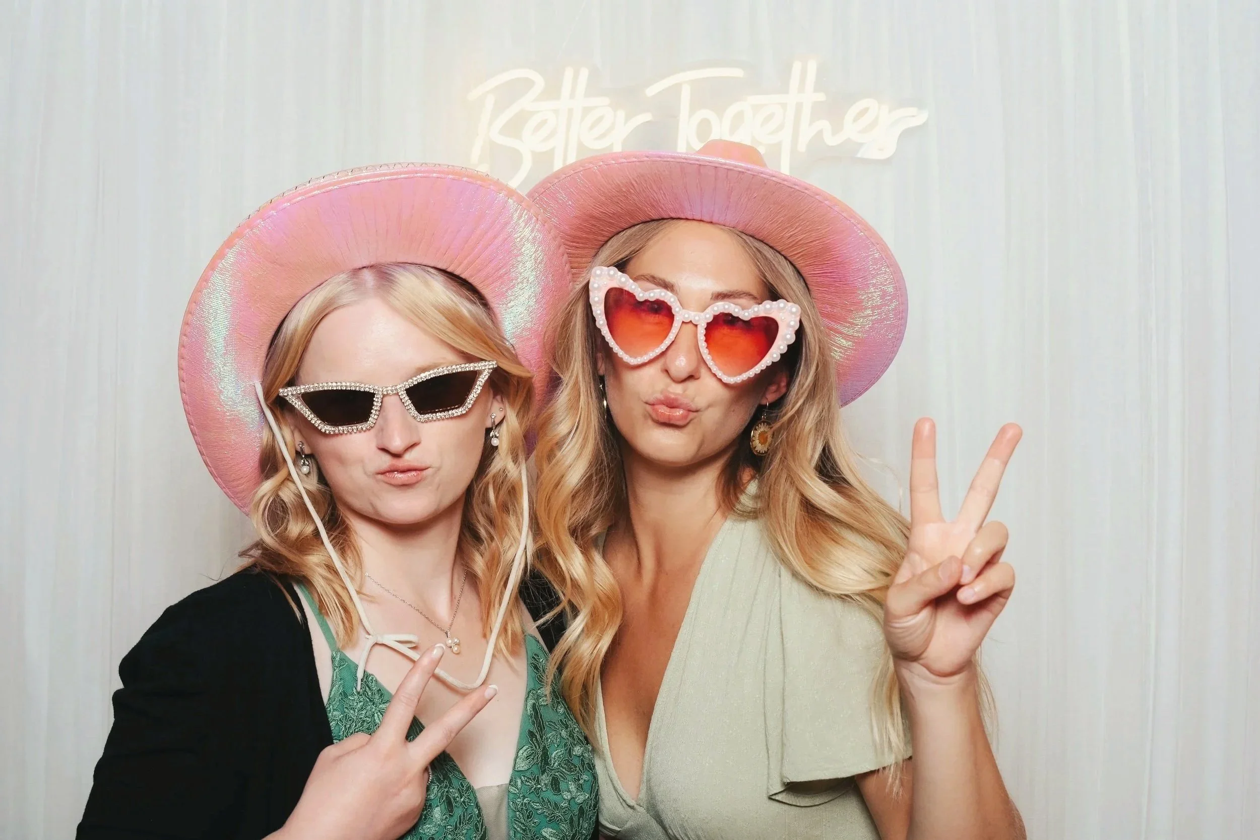 Zwei lachende Frauen mit Sonnenbrillen, Sonnenhüten und Kussmündern posieren in einer Fotokabine. Eine macht ein Peace-Zeichen, im Hintergrund ein Schriftzug 'Better Together'.