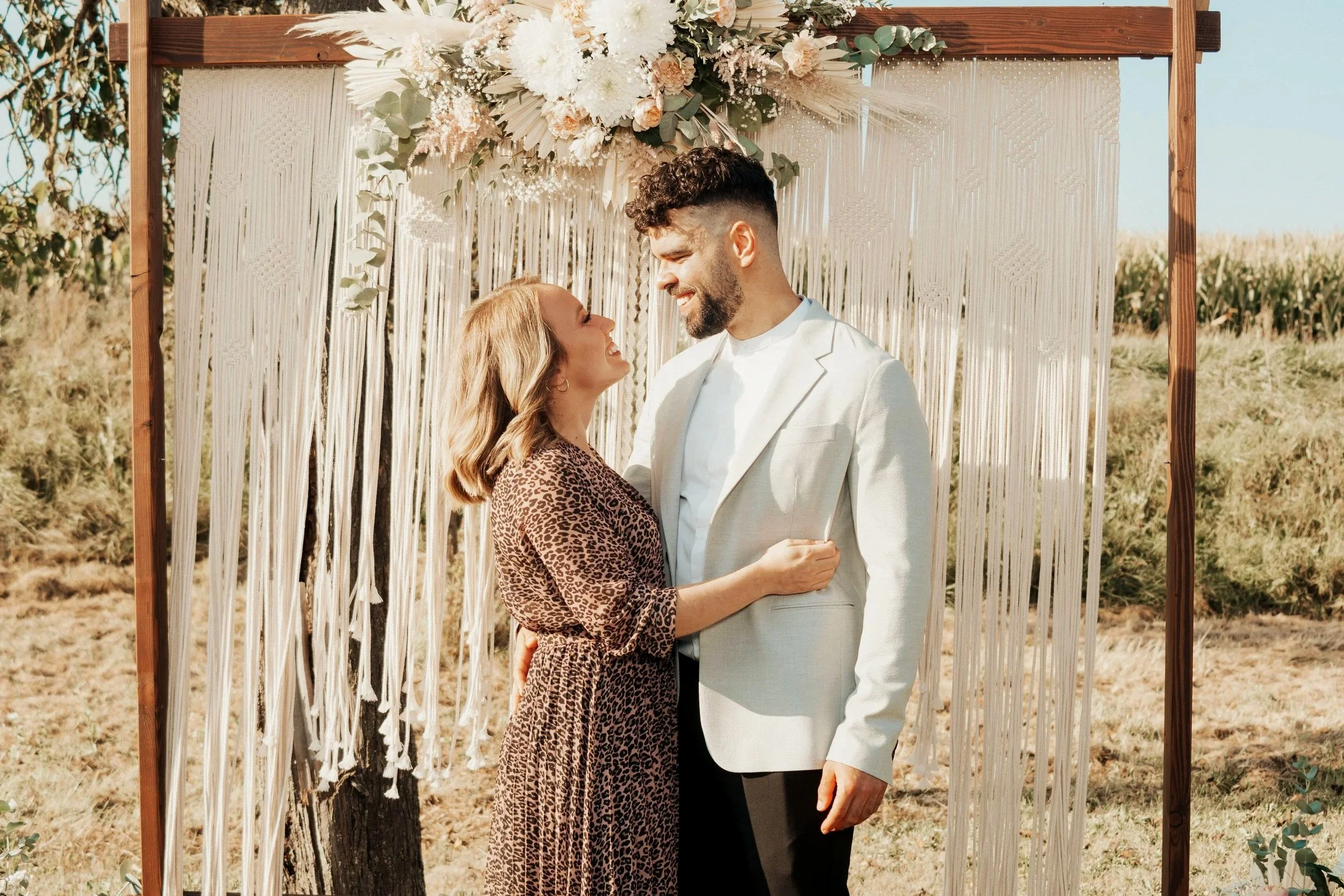 Ein Paar bei einer Hochzeitszeremonie im Freien, vor einem dekorierten Holzrahmen mit Blumen und weißen Stoffen, umarmt und lachend.
