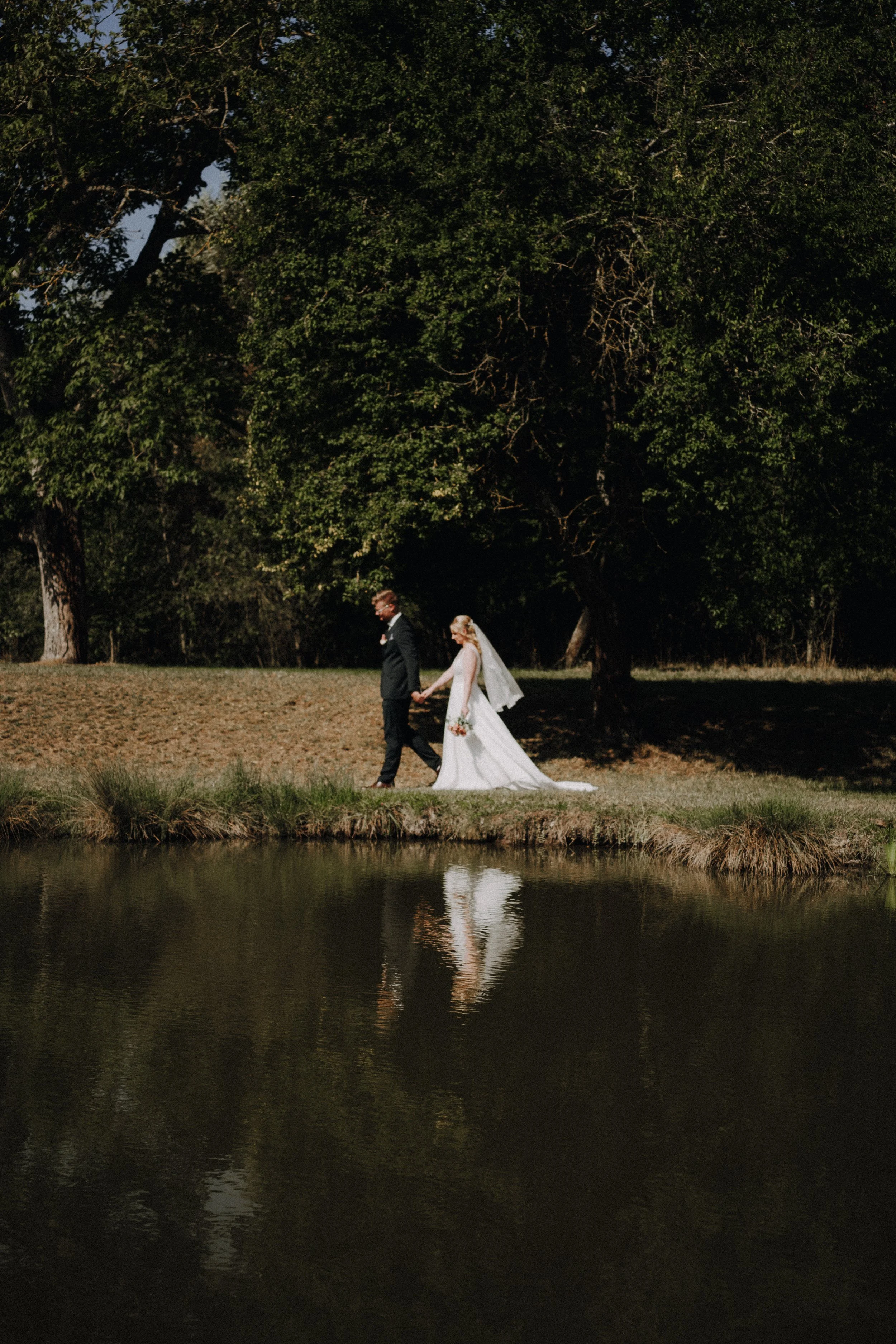 Brautpaar spaziert Hand in Hand an einem See im Freien, im Hintergrund grüne Bäume, bei Sonnenlicht.
