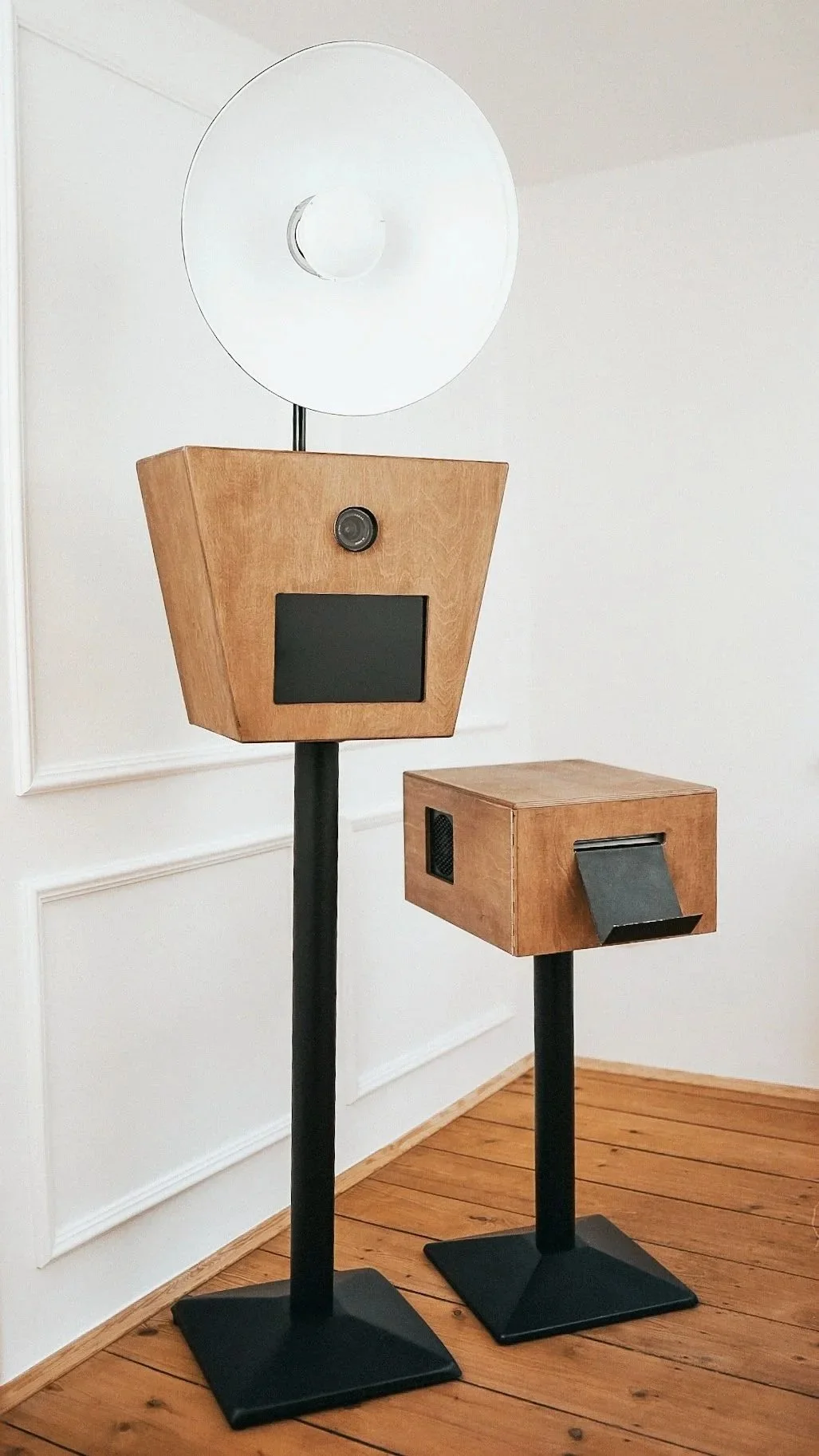 Zwei geometrische Geräte, ein Taller als Balken, ein kurzer und ein langer, mit hölzernen Kuben, einer mit einem Bildschirm und einer Kamera, der andere mit einer Schublade, auf einem Holzfußboden in einem weißen Raum.