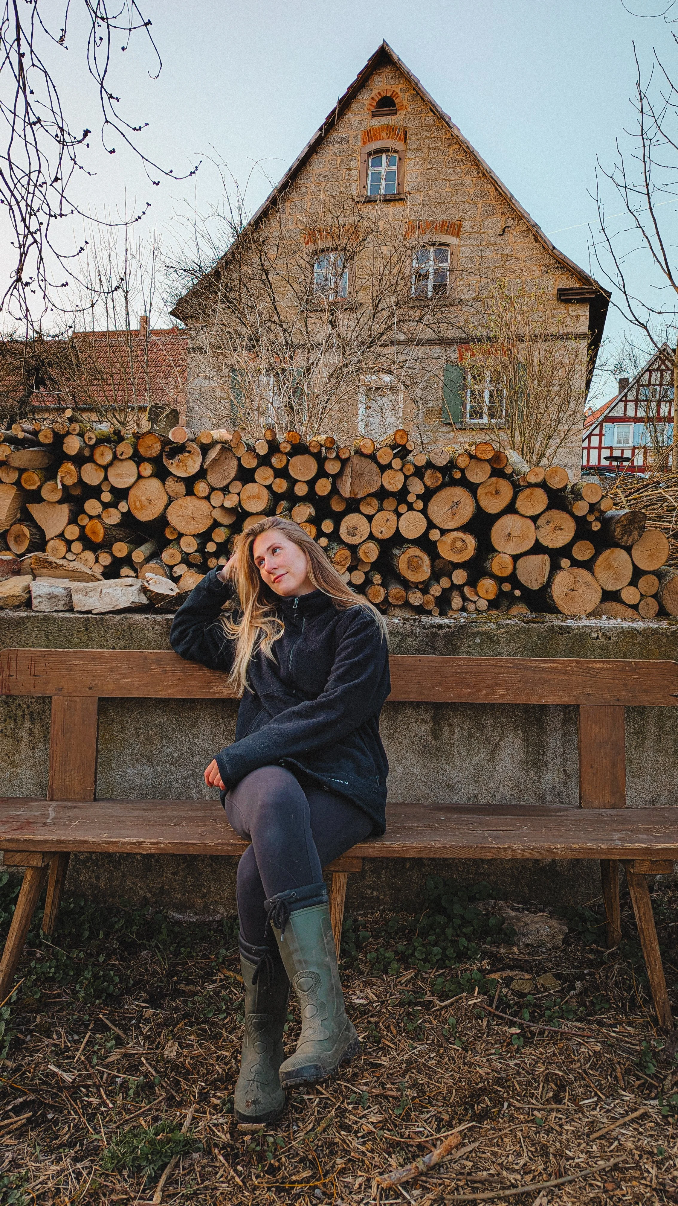 Junge Frau sitzt auf einer Parkbank vor einem Stapel Holzscheite, im Hintergrund ein altes Haus, Blätter an den Bäumen sind kaum sichtbar.