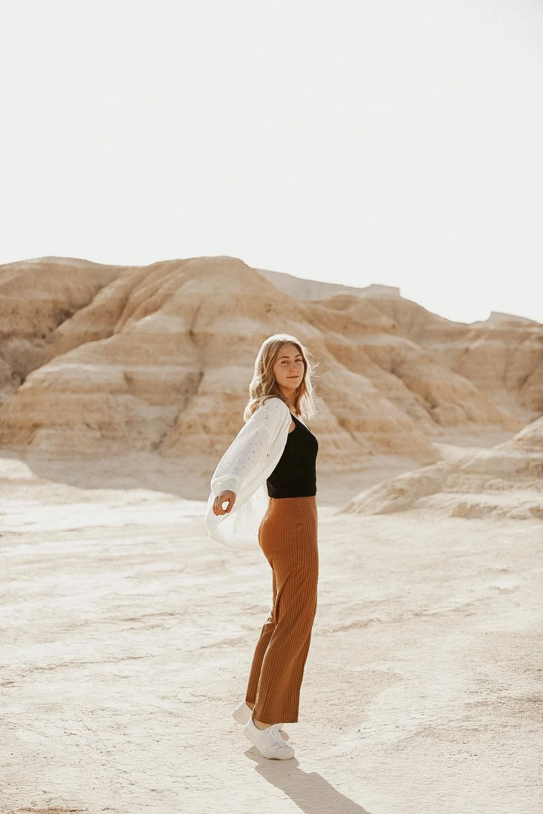 Junge Frau steht in einer Wüste mit sandfarbenen Hügeln im Hintergrund, trägt weiße Sneakers, braune Hosen, ein schwarzes Top und eine weiße Jacke, blickt in die Kamera.
