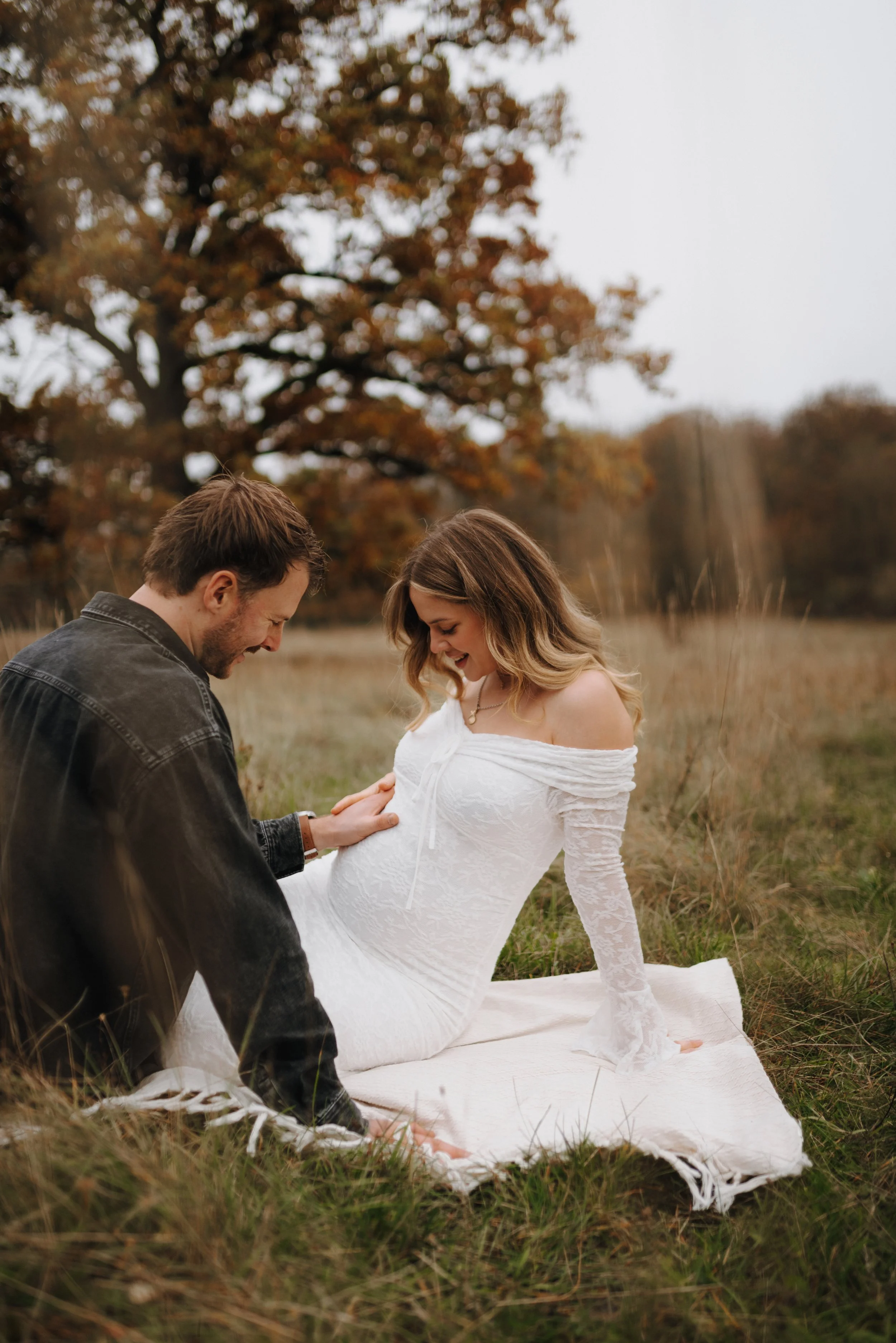 Ein Paar sitzt in einem Herbstwald, die Frau trägt ein weißes, langärmeliges Kleid und der Mann ein dunkles Jackett. Sie scheinen in einem intimen Moment verbunden zu sein.