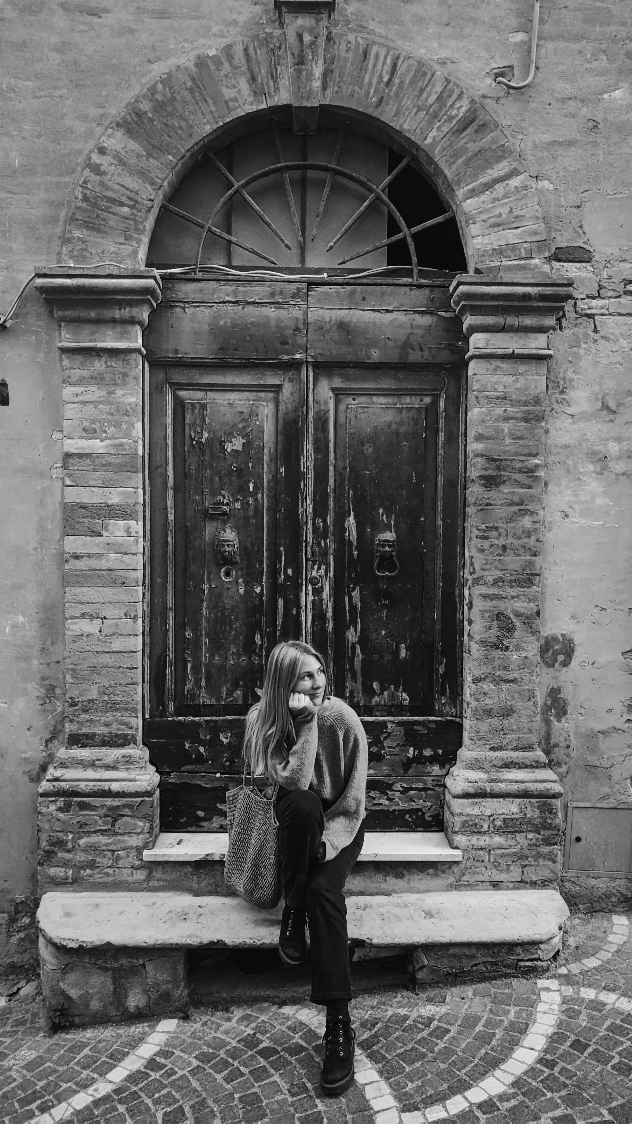 Junge Frau sitzt vor einer alten, holzverschlossenen Tür in einer Steinmauer, umgeben von Ziegeln, mit einem Snack in der Hand und nachdenklich.