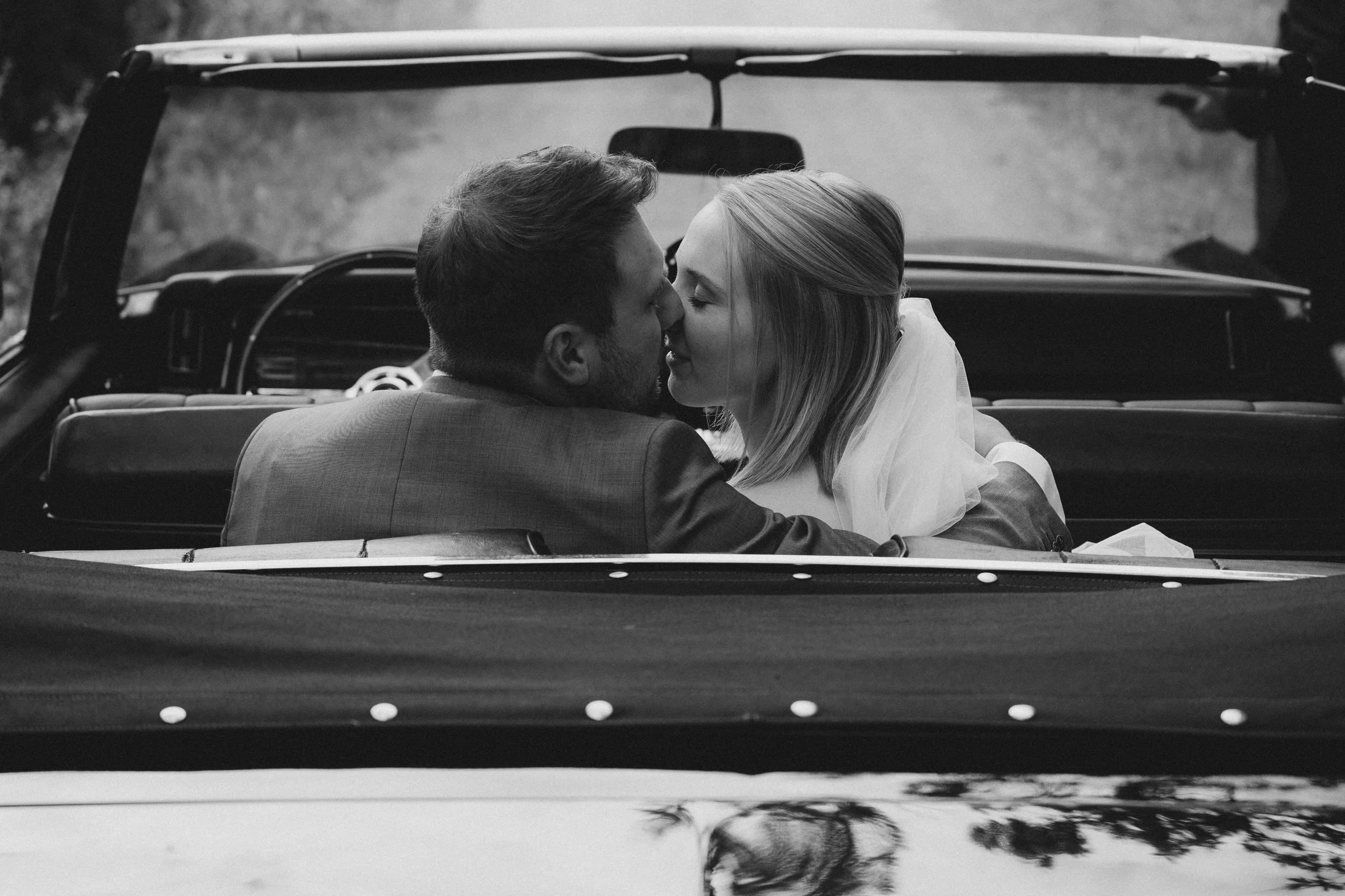 Ein Brautpaar sitzt in einem offenen Auto, küsst sich und ist an einem sonnigen Tag draußen.