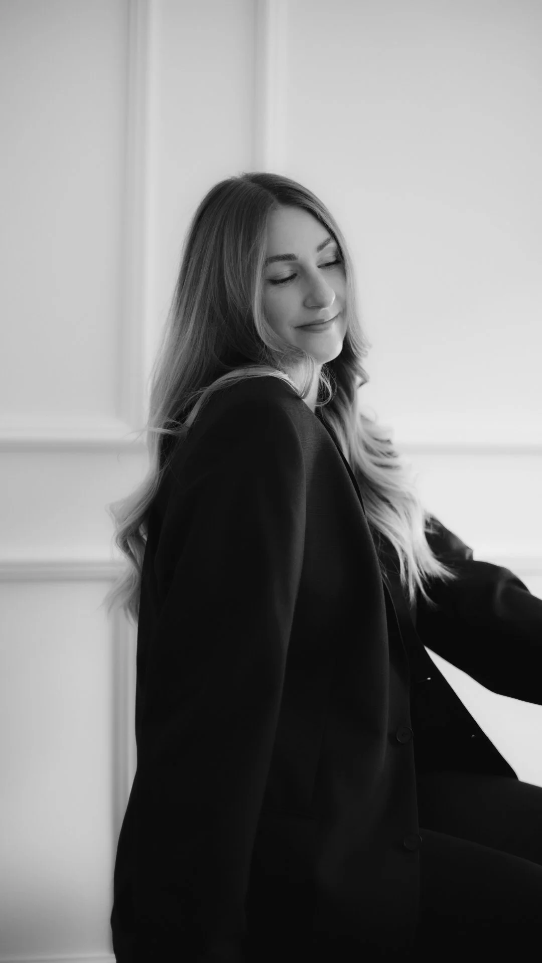 Schwarz-weiße Fotografie einer lächelnden Frau mit geschlossenen Augen, sitzend gegen eine weiße Wand, trägt eine dunkle Jacke.