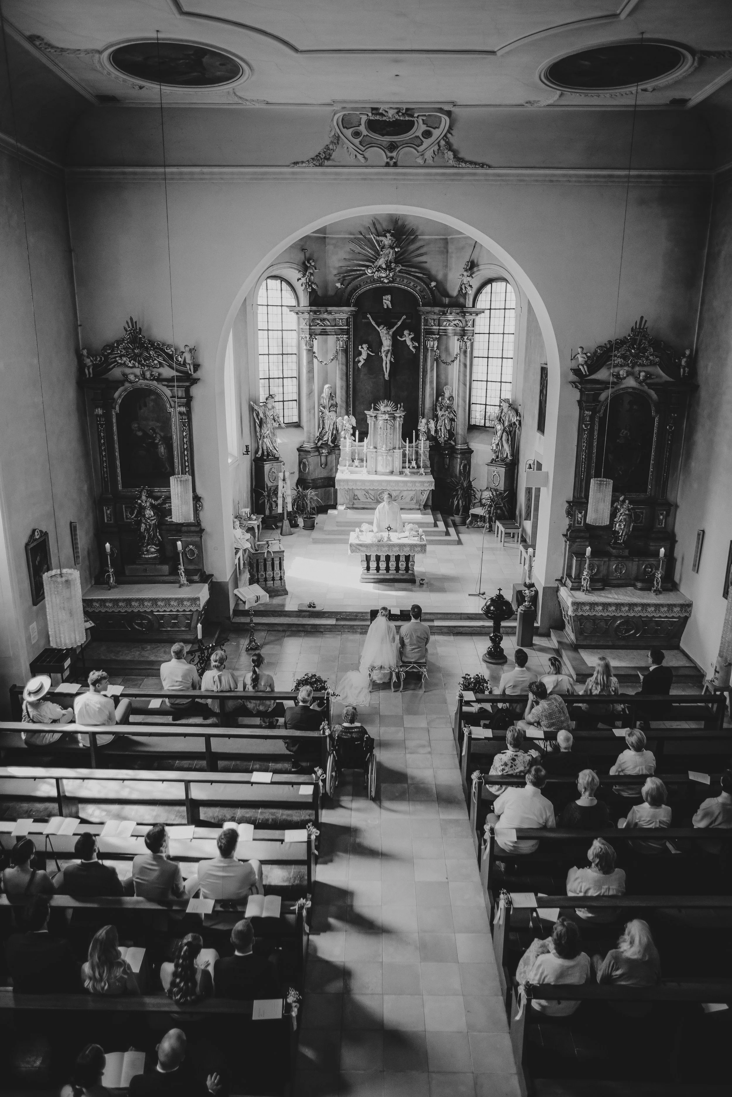 Schwarz-weiß-Foto einer christlichen Hochzeitsmesse in einer kleinen Kirche, mit Brautpaar vor dem Altar, umgeben von Gottesdienstbesuchern, die auf Bänken sitzen. Der Altar ist mit religiösen Statuen und Dekorationen geschmückt.