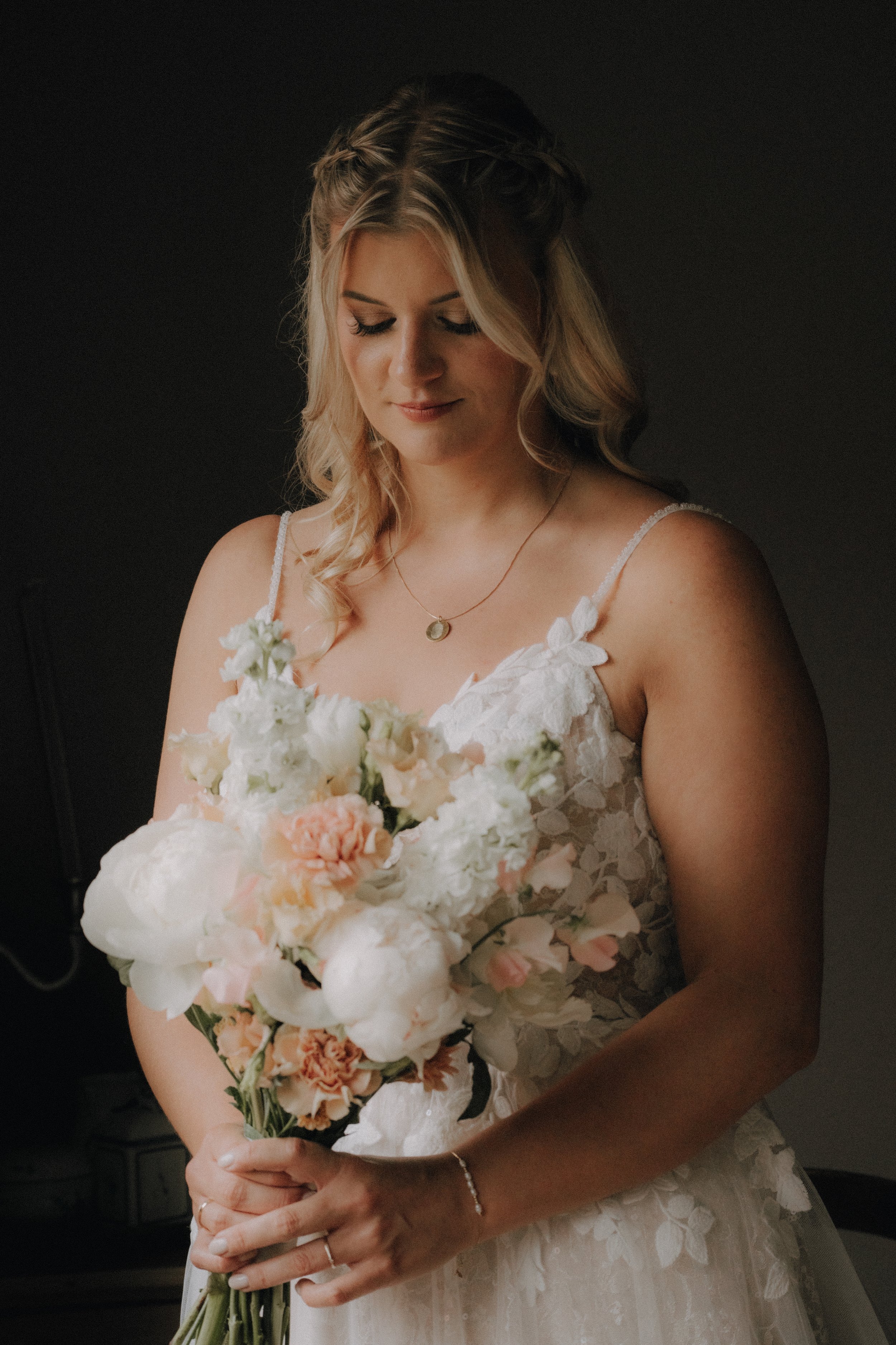 Braut in einem weißen Hochzeitskleid mit Blumenmuster hält einen Blumenstrauß mit weißen und hellrosanen Blumen, hat blonde Locken, trägt eine goldene Halskette und ein Armband, steht vor einem dunklen Hintergrund.