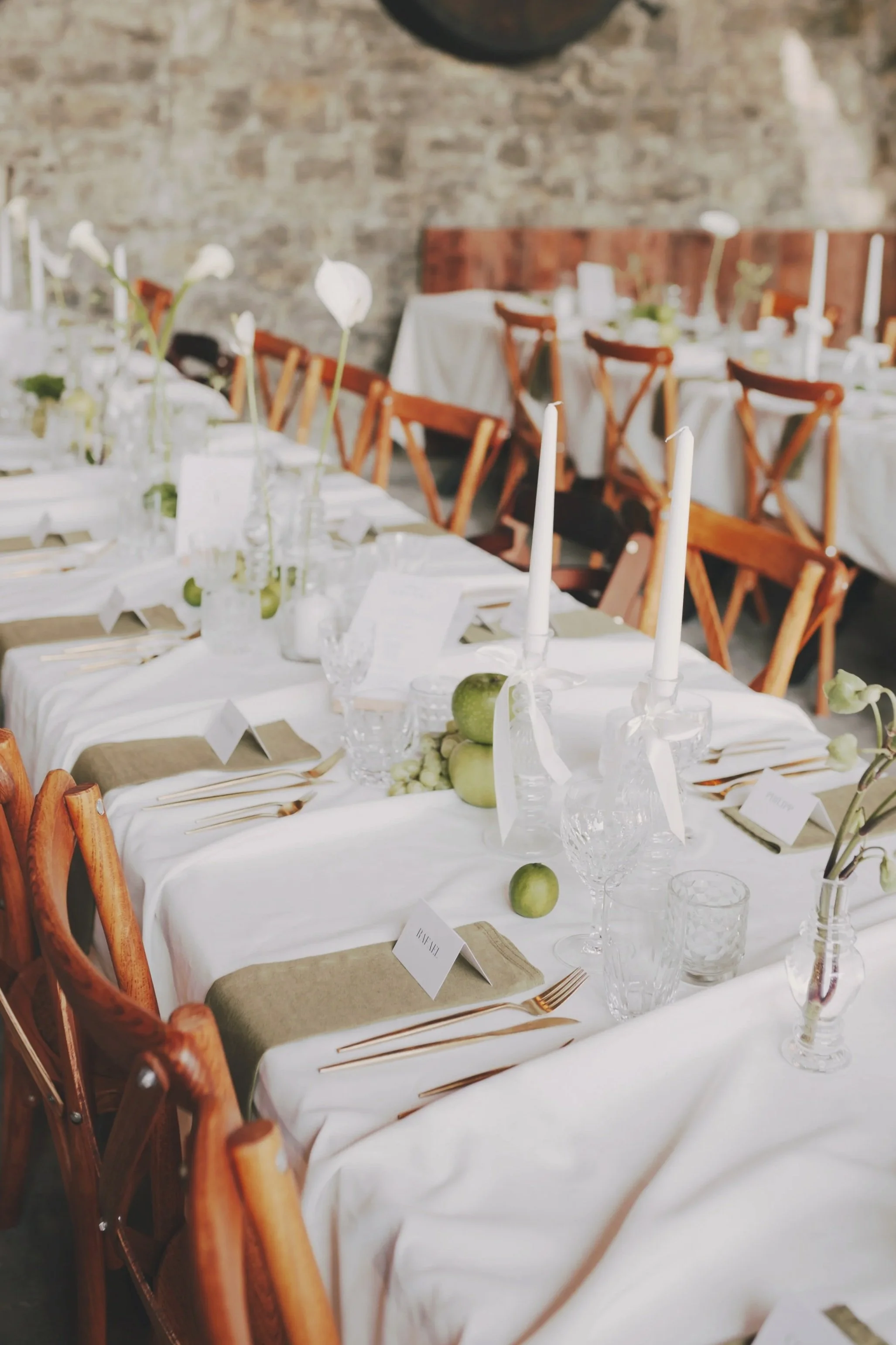 Ein festlich gedeckter Esstisch mit weißen Tischdecken, Kerzen in Kristallleuchtern, grünen Äpfeln und einfachen Blumendekorationen in Gläsern, in einem rustikalen Raum mit Steinwänden.