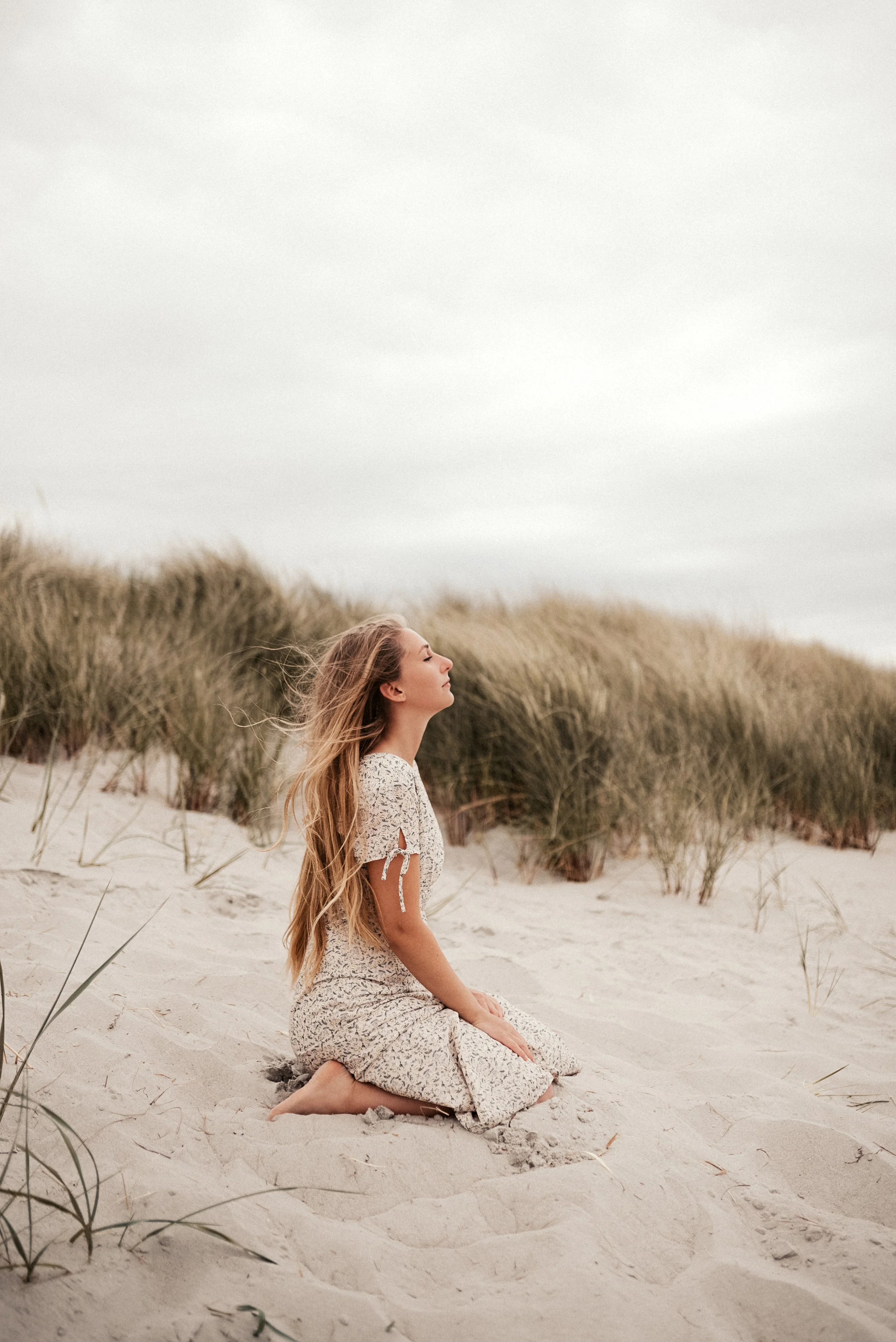 Frau kniet im Sand an Dünen, trägt ein langes Kleid mit Kurzarm, die Augen geschlossen, unter wolkigem Himmel.
