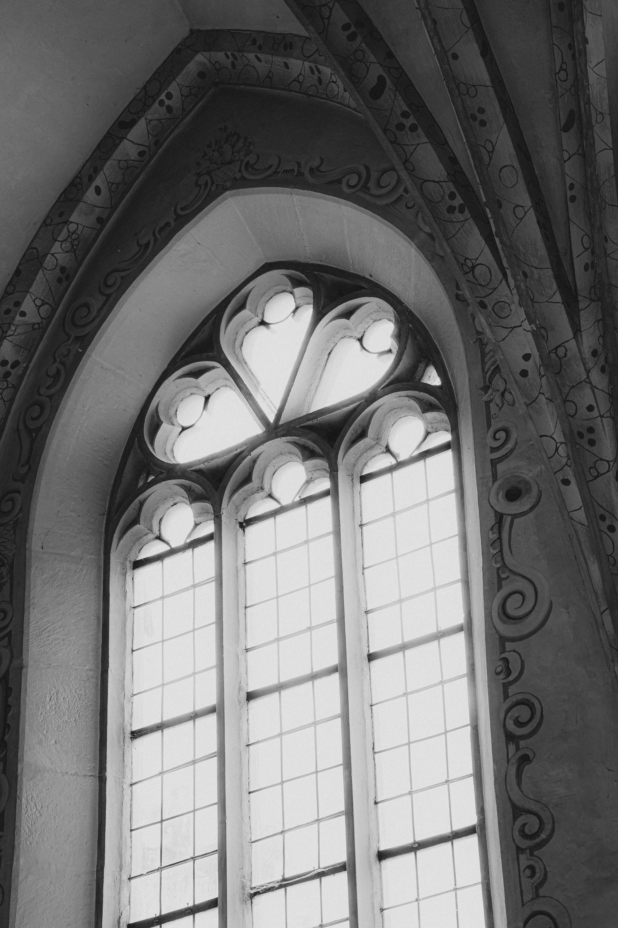 Schwarz-Weiß-Fotografie eines gotischen Kirchenfensters mit kunstvoller, dekorativer Verzierung um den Rahmen.