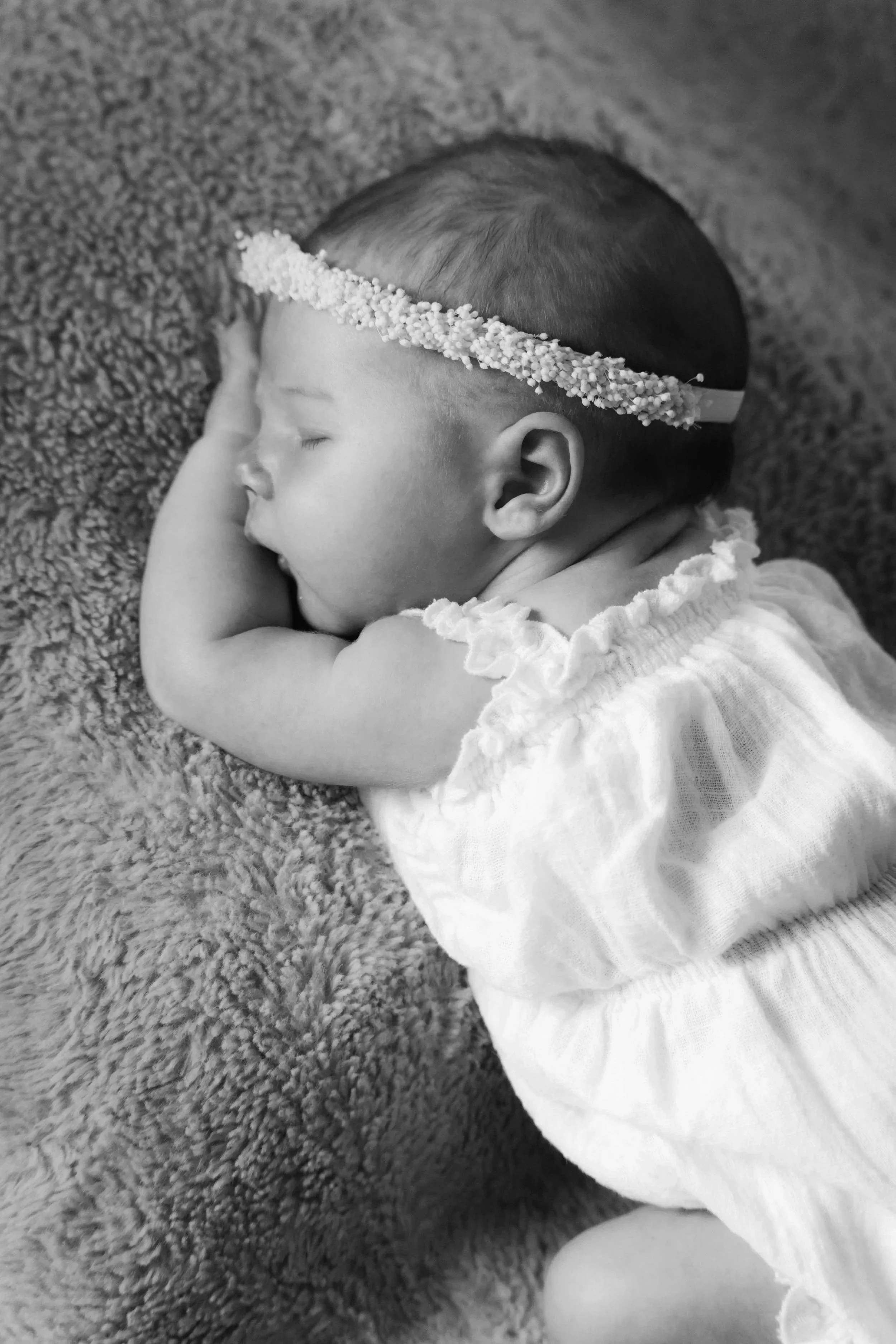 Ein neugeborenes Mädchen schläft auf einem weichen, flauschigen Kissen, trägt ein lockeres, weißes Kleid und eine Kopfbedeckung aus kleinen Perlen.