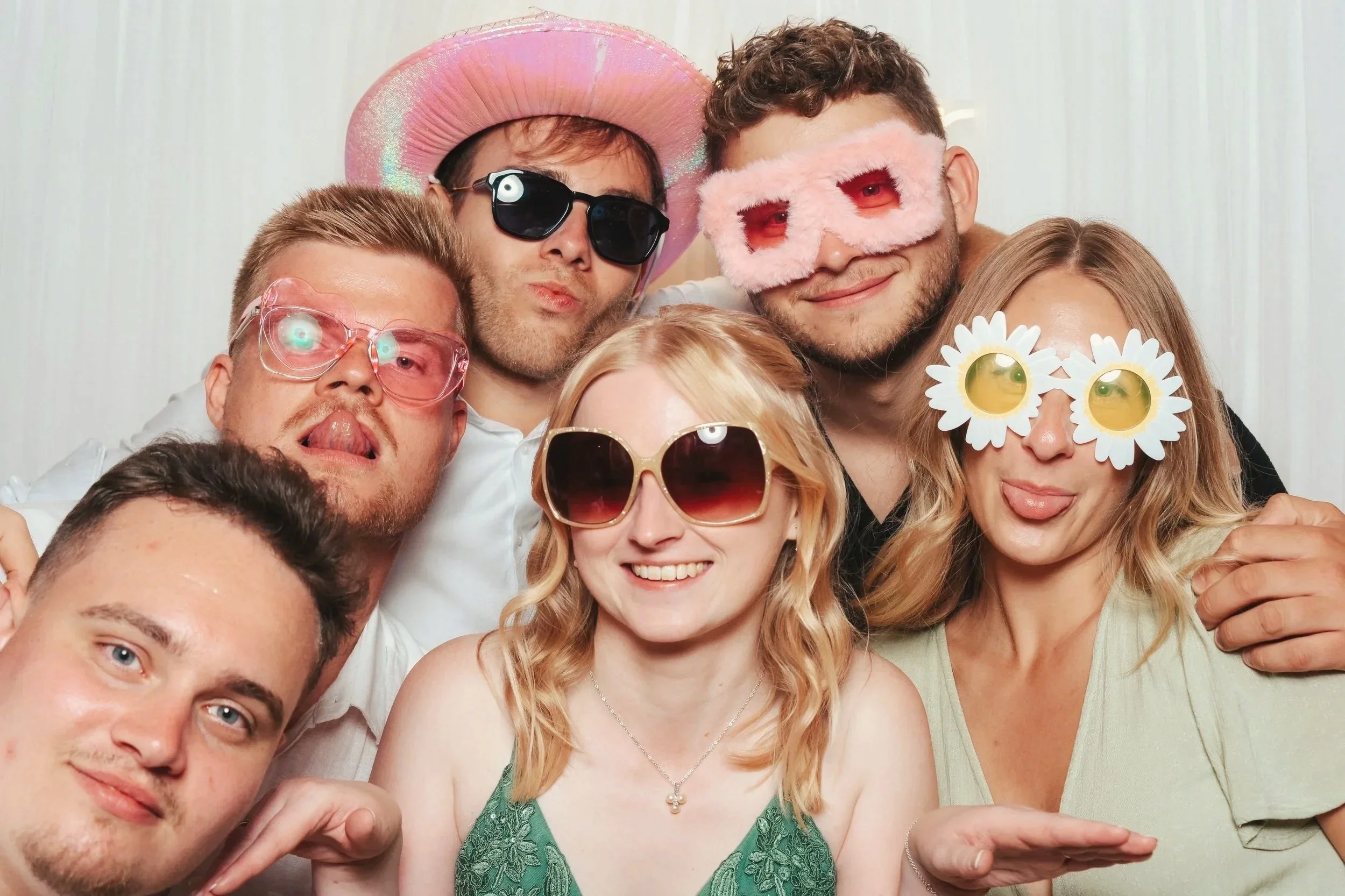 Eine Gruppe von sieben Freunden, die auf einer Party Spaß haben, trägt auffällige und lustige Brillen, Hüte und Masken, posieren fröhlich vor einer hellen, neutralen Hintergrundwand.