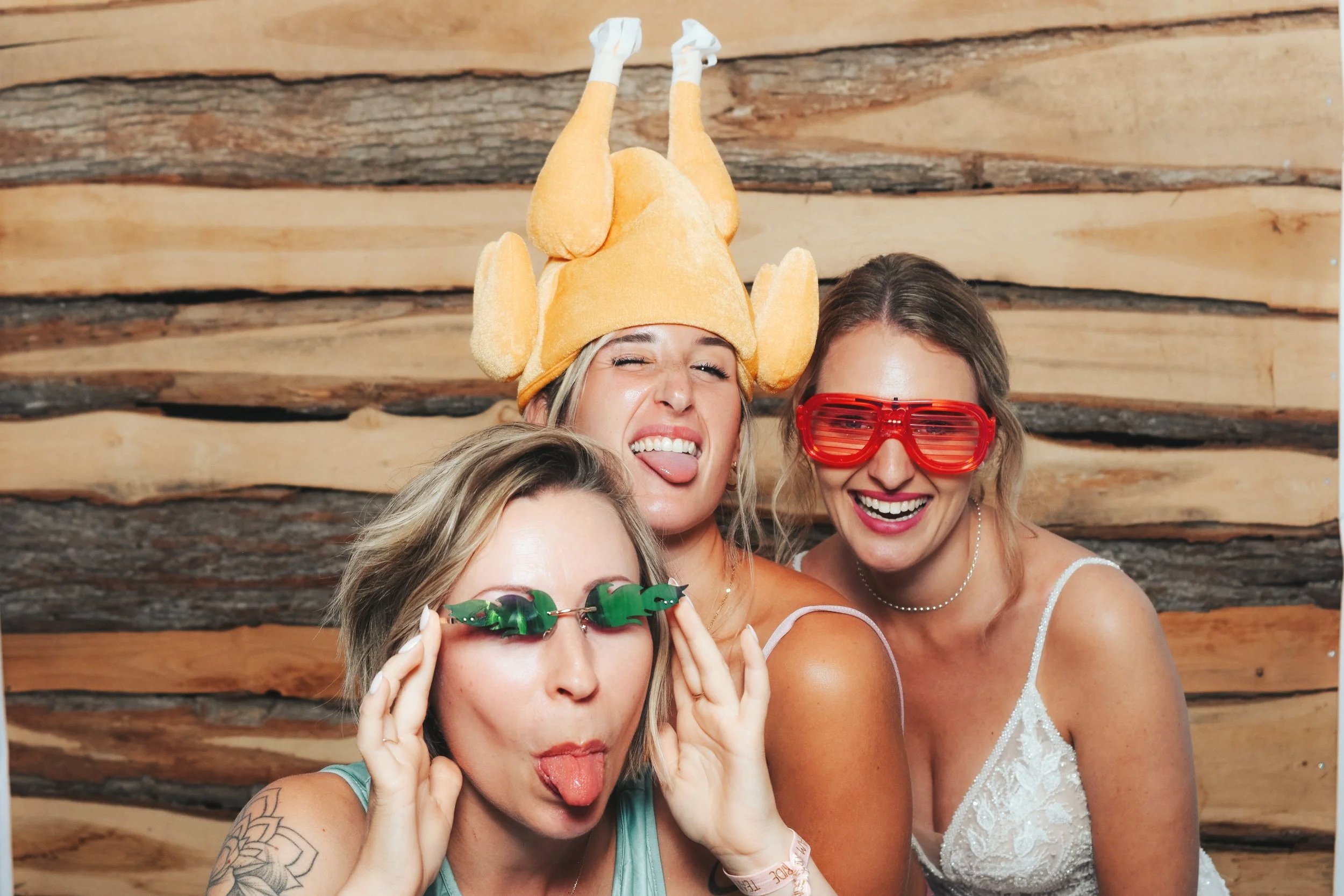 Drei lachende Frauen in Partyoutfits und lustigen Accessoires vor Holz-Wand.