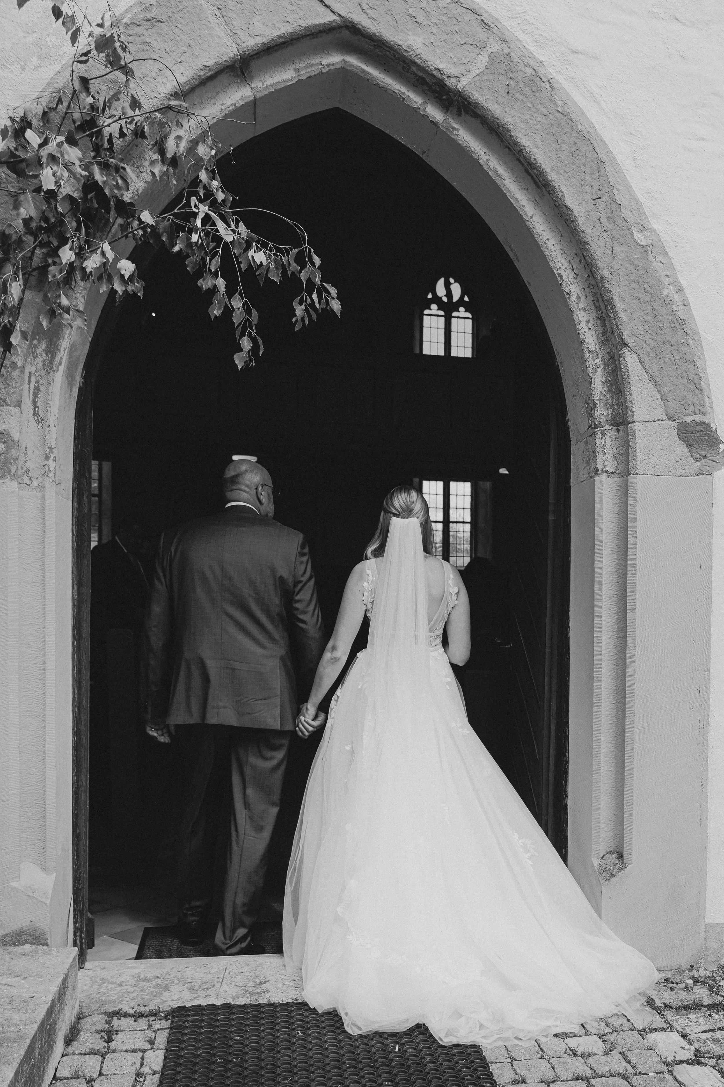 Braut in Hochzeitskleid und Bräutigam in Anzug betreten eine Kirche durch ein Tor.