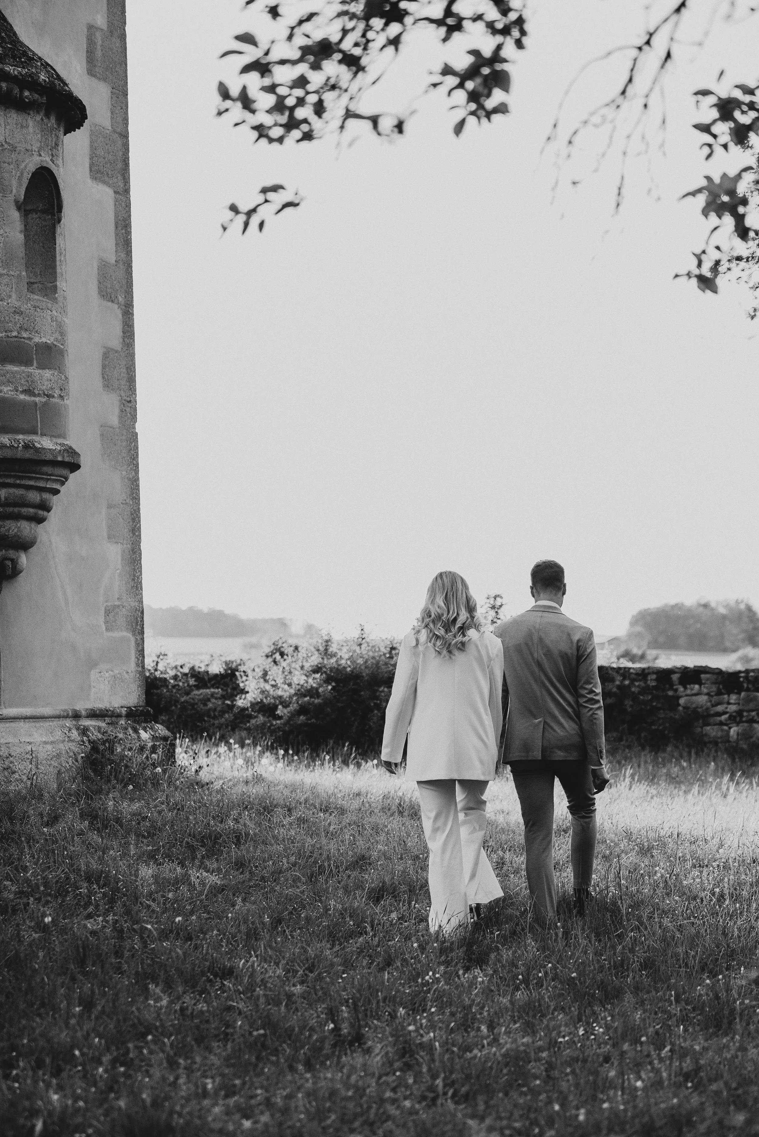 Zwei Menschen, die einen Spaziergang im Freien machen, sehen von hinten. Es ist schwarz-weiß, die Person auf der linken trägt einen hellen Anzug, die Person auf der rechten einen dunklen Anzug. Sie gehen an einer alten Steingebäuden vorbei, im Hintergrund sind Bäume und ein Wasser, wahrscheinlich ein Fluss oder See.