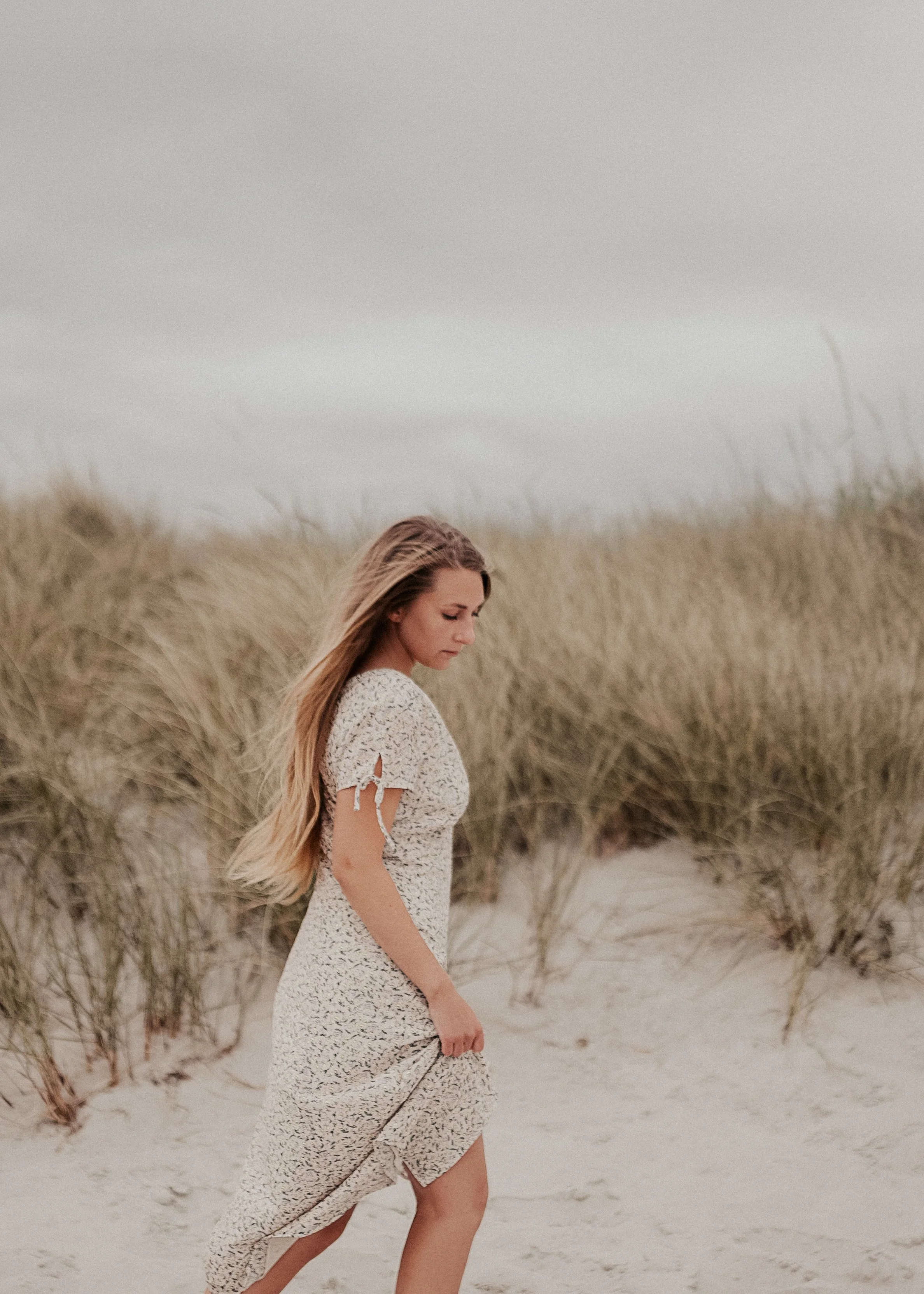 Eine junge Frau in einem weißen Kleid läuft an einer Düne mit hohen Gras auf einem Strand. Der Himmel ist bedeckt und die Stimmung ist ruhig und nachdenklich.