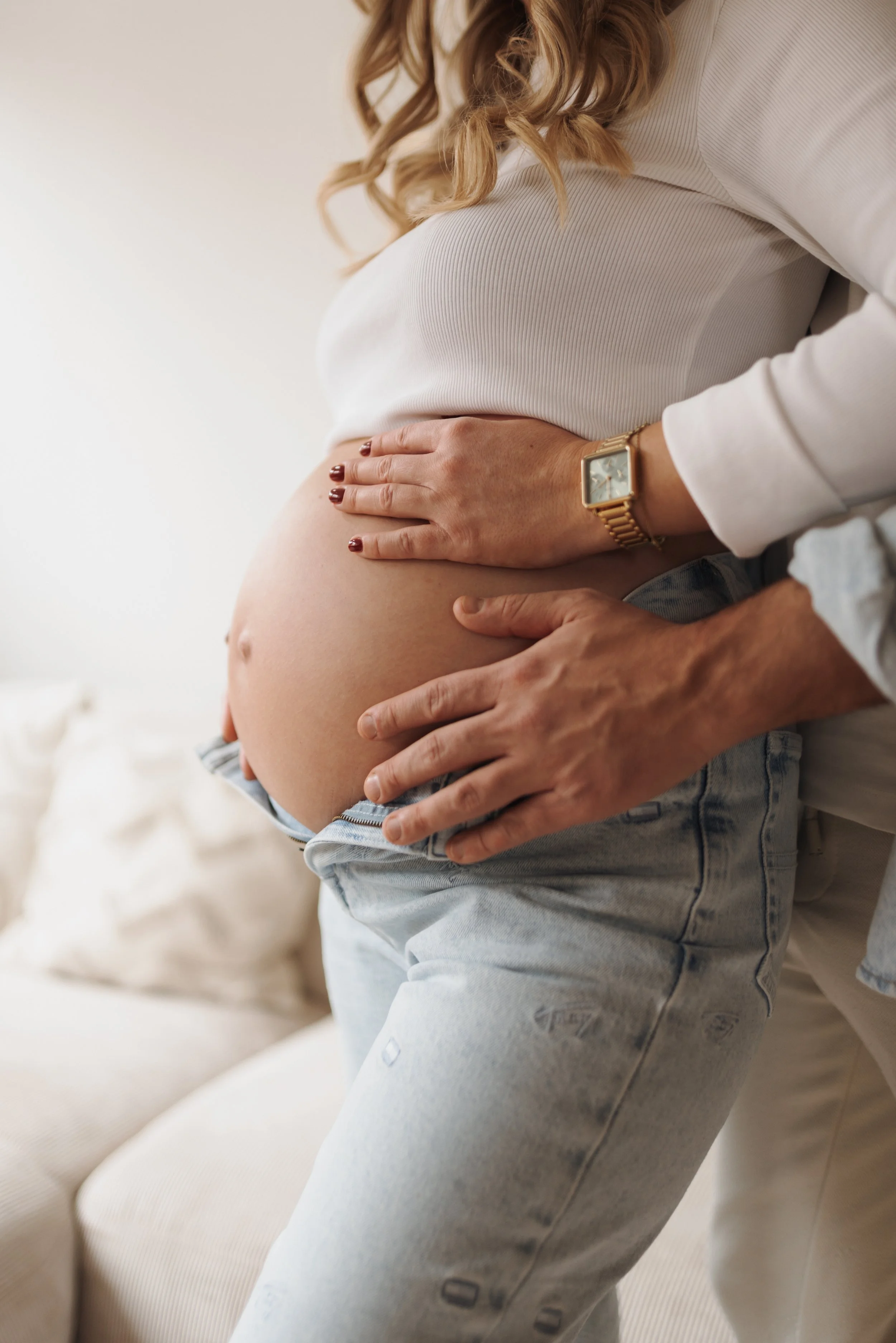 Nahaufnahme eines schwangeren Bauches, gehalten von zwei Personen mit den Händen, im Innenraum mit Sofa und Kissen im Hintergrund.