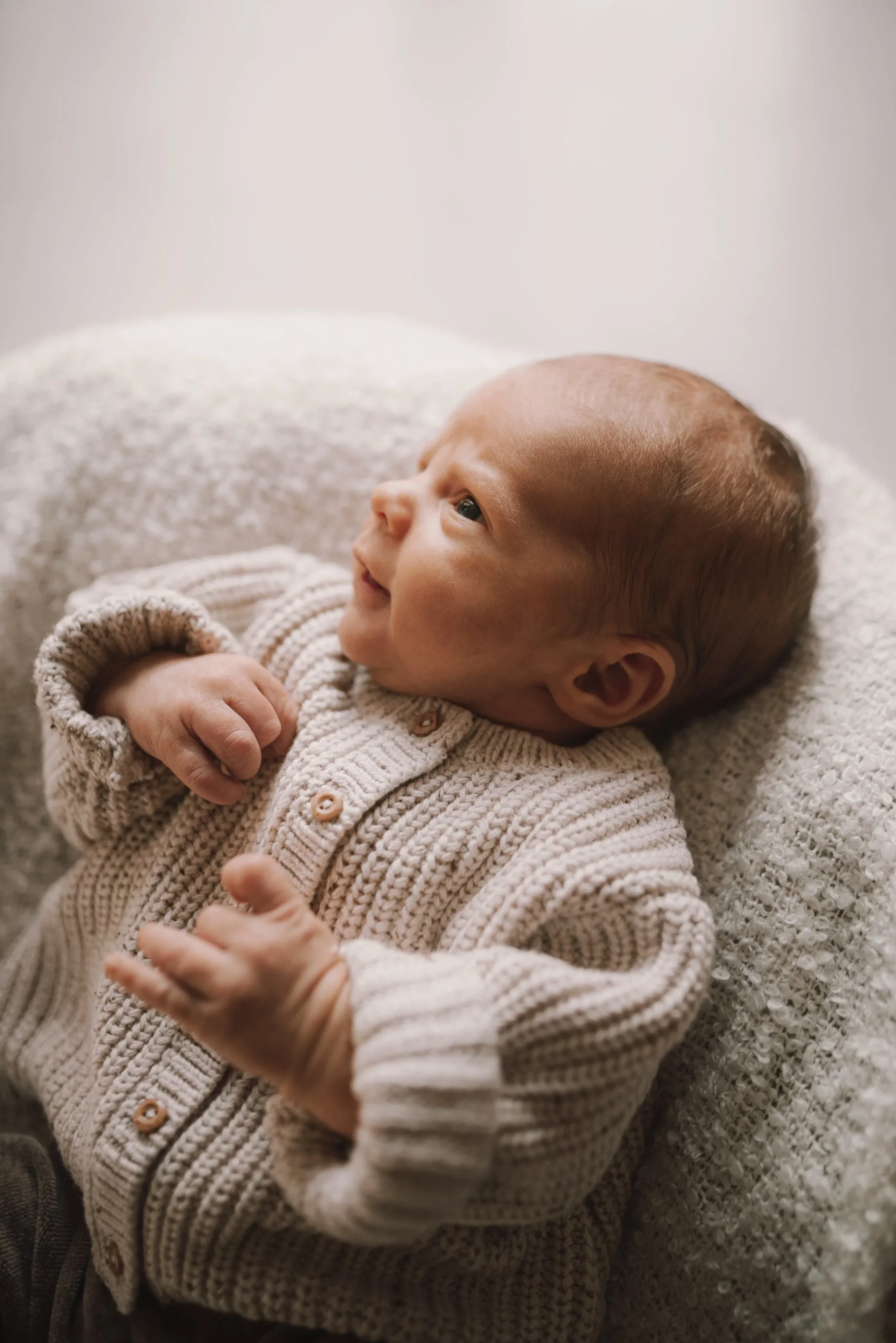 Ein Baby liegt auf einem weichen, cremefarbenen Kissen, trägt einen beigefarbenen Strickpullover und schaut neugierig nach oben.