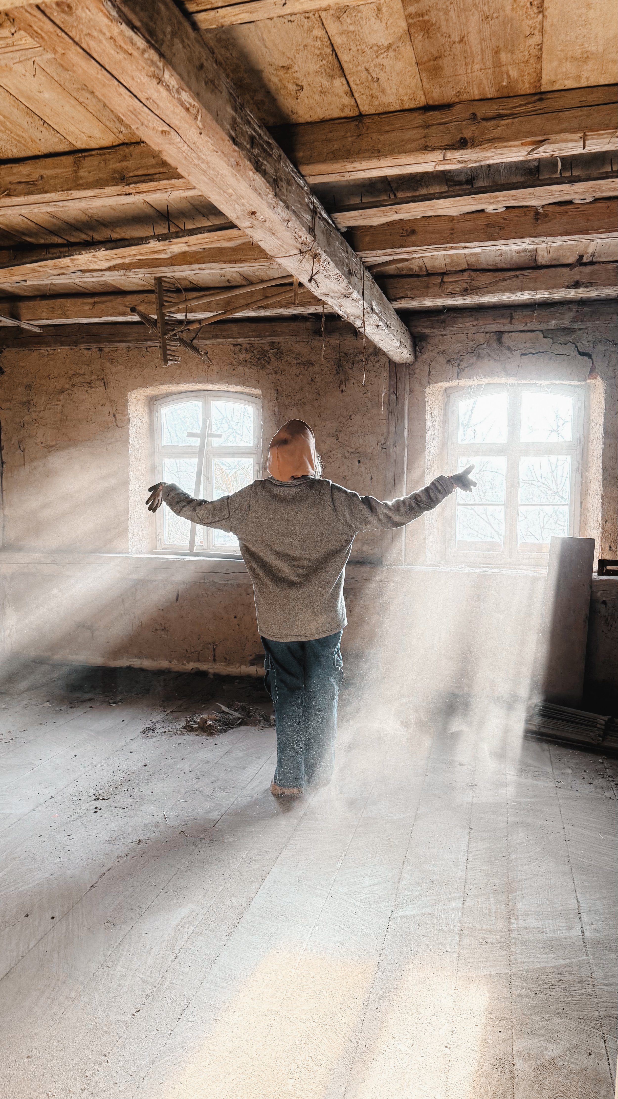 Eine Person steht in einem unvollendeten, alten Raum mit Holzbalken, Sonnenlicht durch zwei Fenster, Staub in der Luft, mit ausgestreckten Armen, die Rückenansicht.