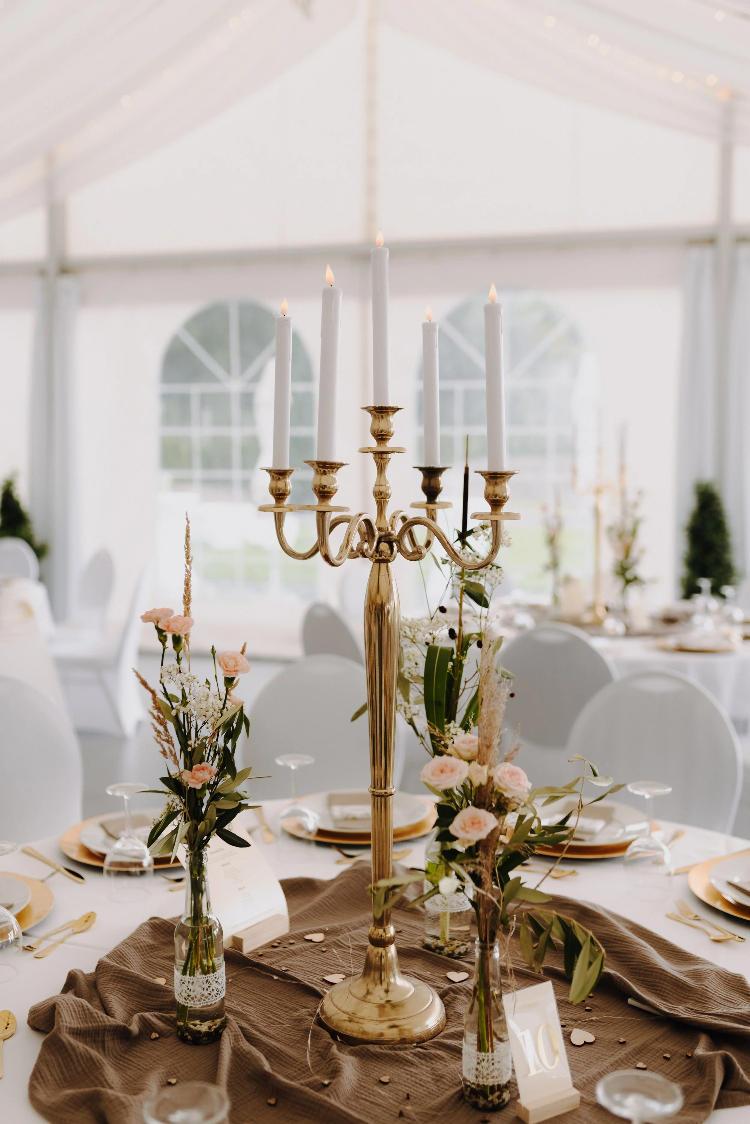 Eleganter festlicher Tisch mit goldener Kerzenhalter, weißen Kerzen, Blumenarrangements und goldenen Servietten in einer großen weißen Zeltveranstaltung.