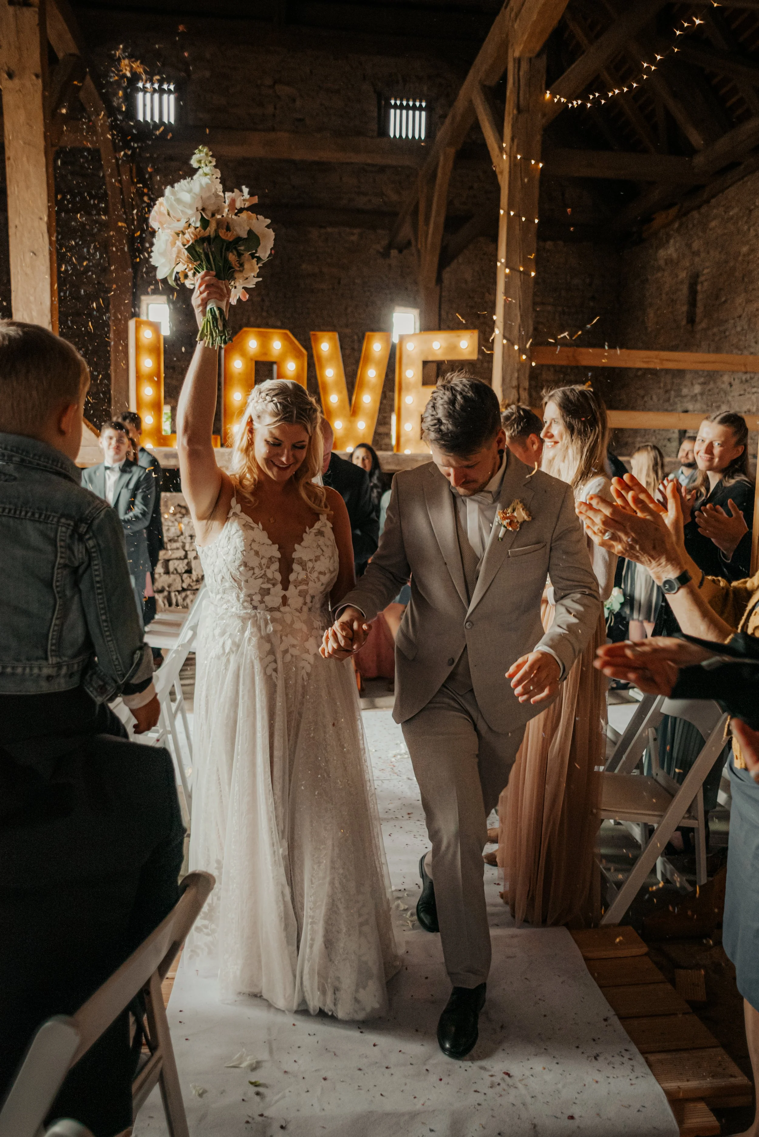 Ein Brautpaar bei ihrer Hochzeit, die Braut hält einen Blumenstrauß hoch, während sie durch einen Gästenapplaus geht, im Hintergrund leuchtende Buchstaben, die 'LOVE' schreiben.