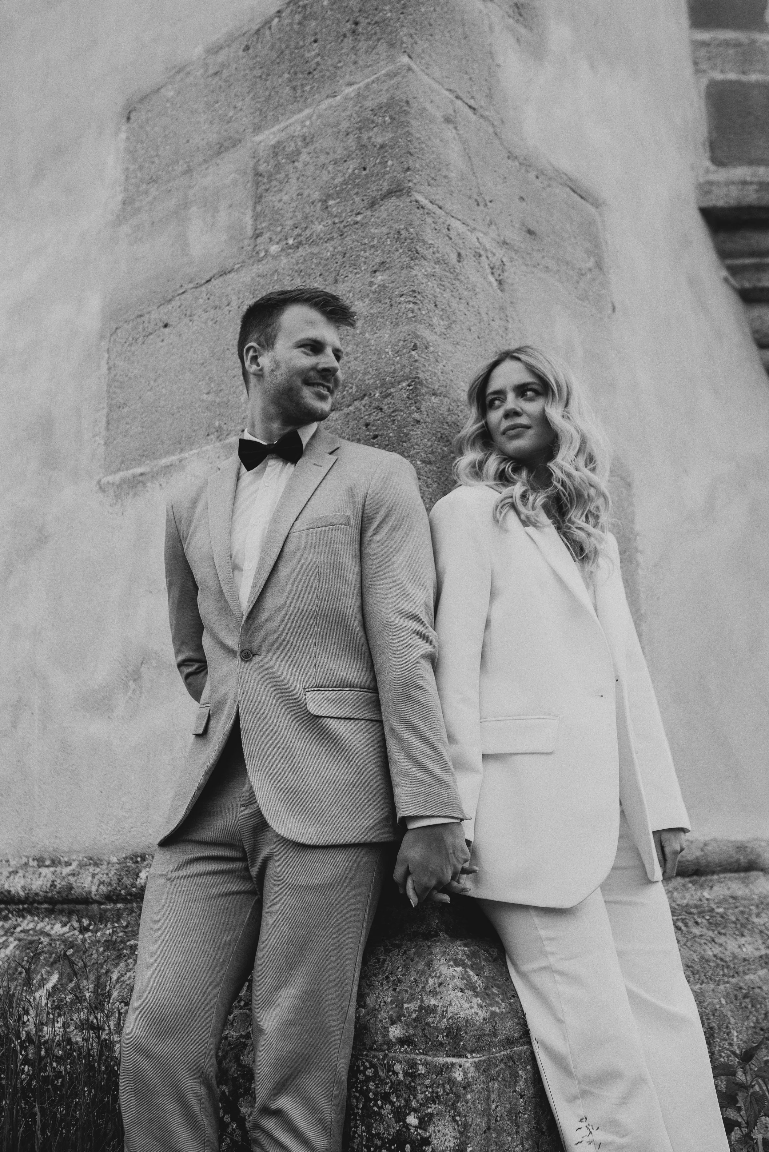 Ein Mann in einem Anzug mit Fliege und eine Frau in einem weißen Anzug stehen nebeneinander und halten sich an den Händen, vor einer alten Steinmauer, schwarz-weiß Fotografie.