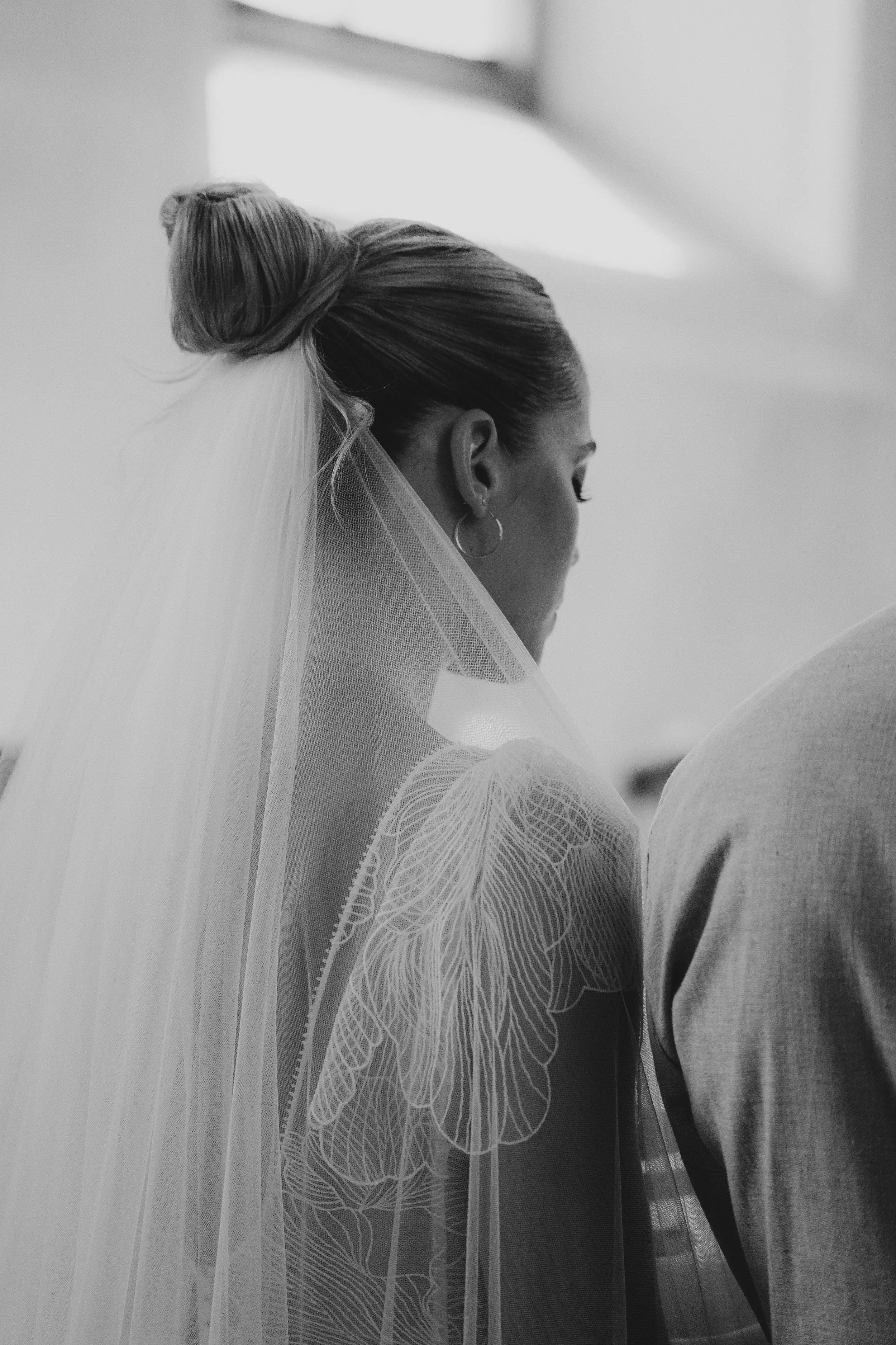 Schwarze und weiße Nahaufnahme einer braut mit hochgestecktem Haar, einer langen Tüllveil, die mit Spitzenmustern verziert ist, und einem weißen Brautkleid. Sie steht neben einem Mann, vermutlich dem Bräutigam, bei einer Hochzeit.