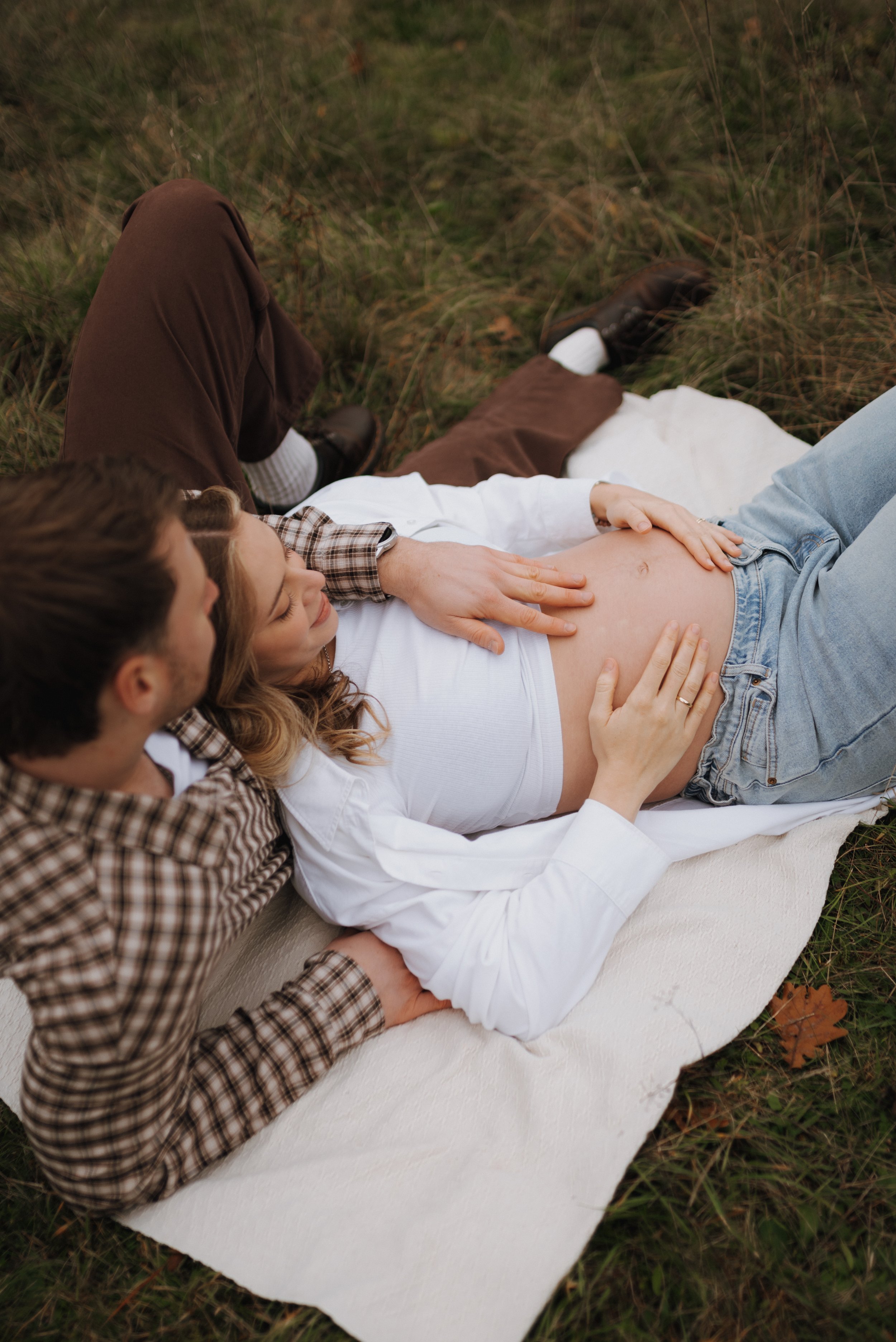 Paare liegen auf einer Matte im Freien, wobei die schwangere Frau ihre Bauch berührt und der Mann ihre Hand hält.