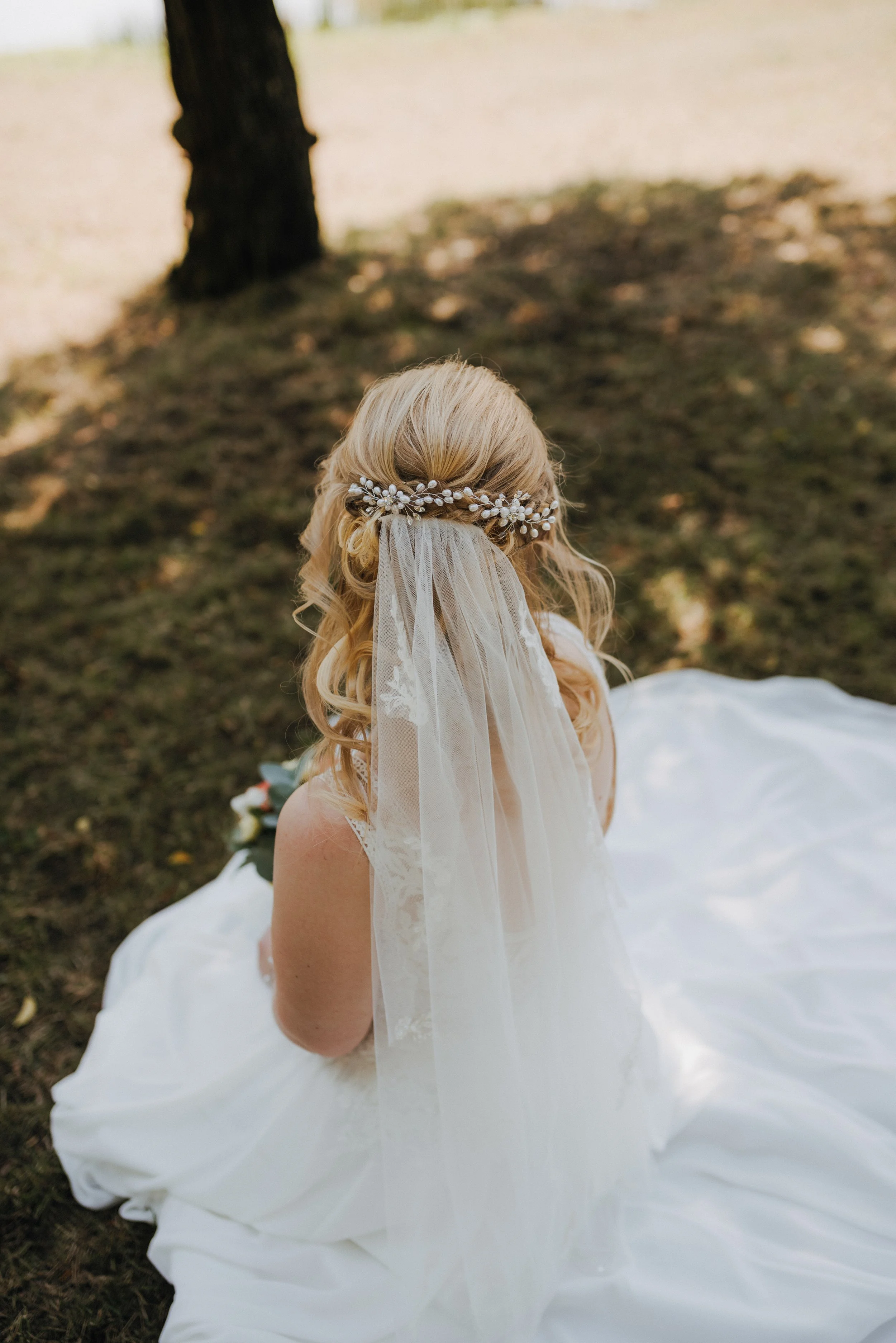 Braut mit langem weißen Kleid, mit Blumenkranz und Schleier, sitzt im Freien auf dem Boden, von hinten sichtbar, mit einem Baum im Hintergrund.