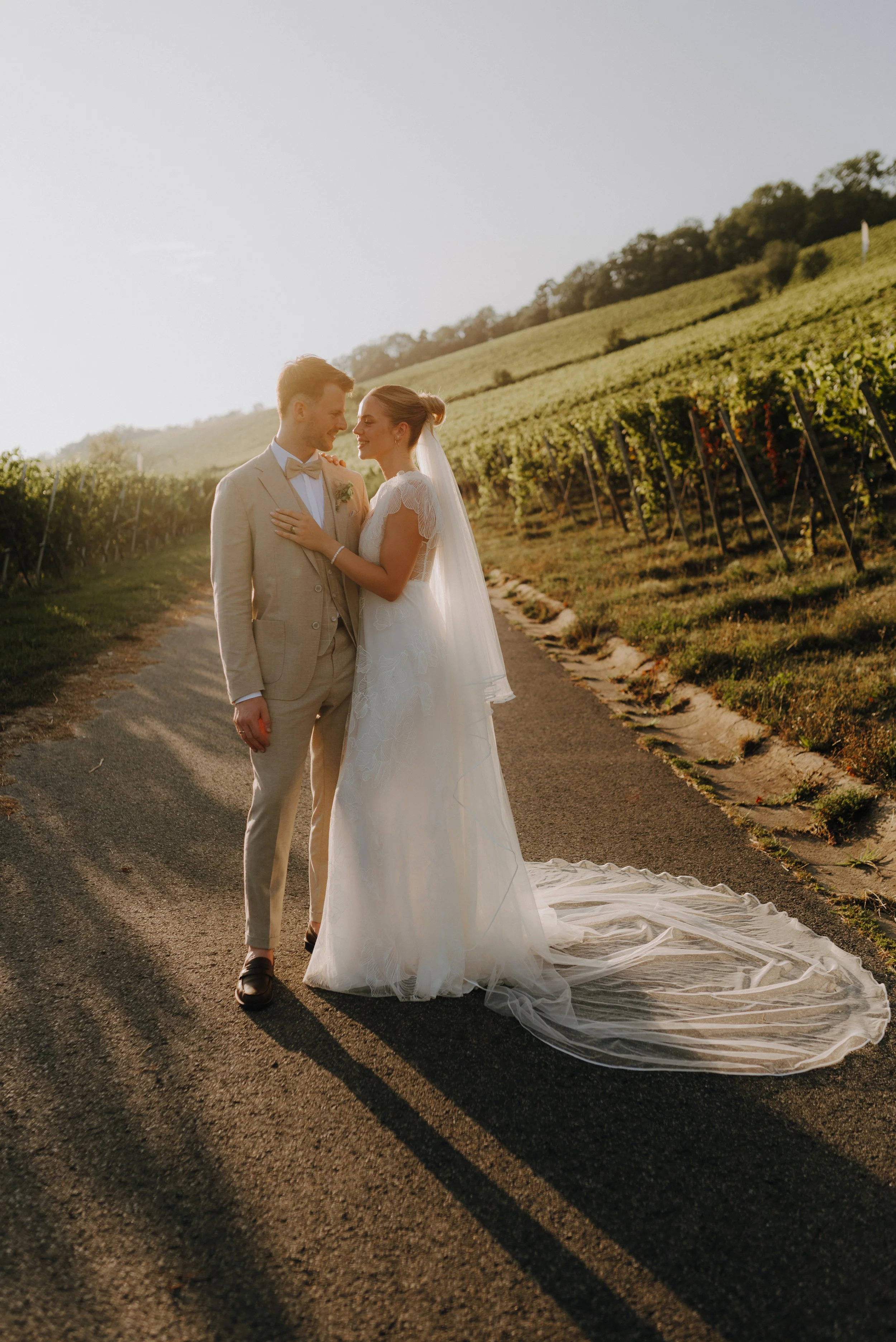 Ein Brautpaar in Hochzeitskleidung steht sich liebevoll auf einer Landstraße bei Sonnenuntergang gegenüber. Der Hintergrund zeigt Weinberge und einen bewölkten Himmel.
