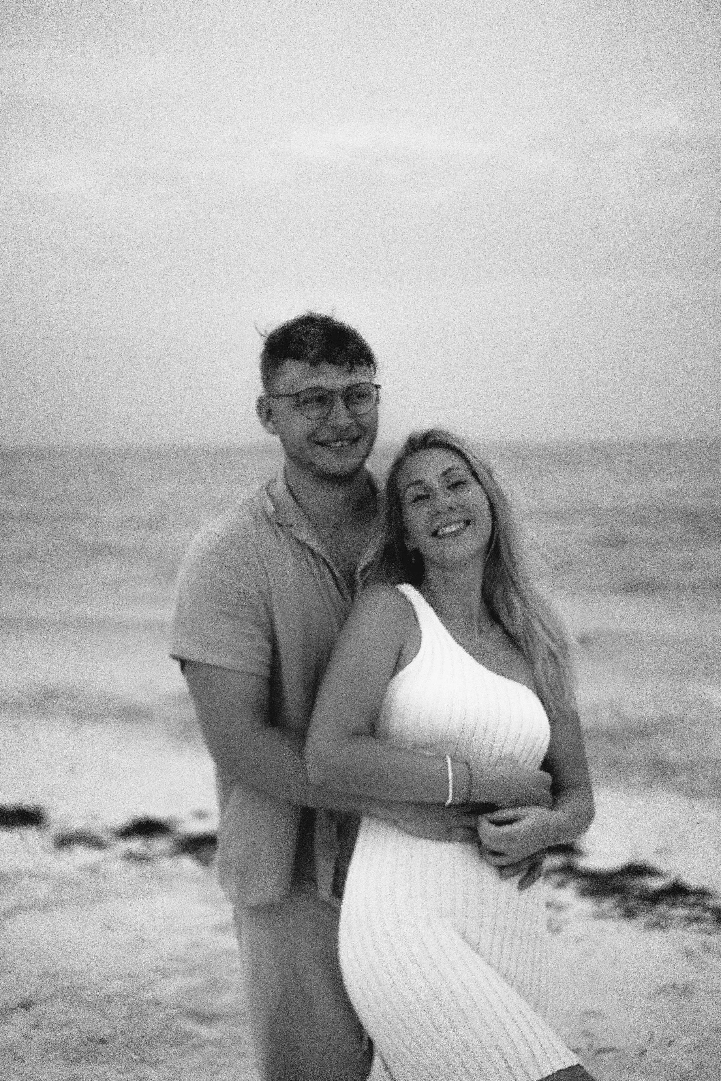 Ein lächelndes Paar steht am Strand mit Meer im Hintergrund, schwarz-weiß Fotografie.