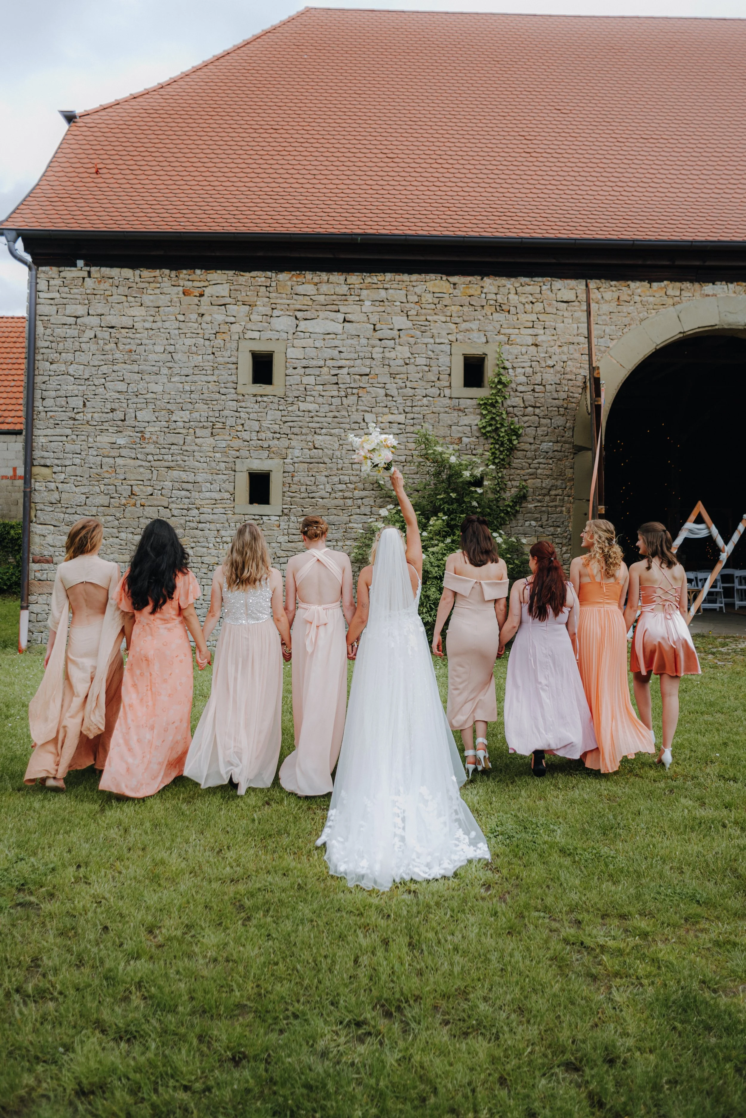 Hochzeitspaar mit Braut im weißen Kleid und Brautjungfern in pastellfarbenen Kleidern, läuft auf grünem Rasen vor einer alten Steinmauer mit Tür erwartet, die in einem offenen Veranstaltungsort führt.