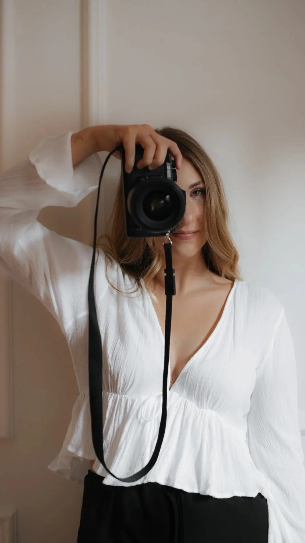 Eine junge Frau macht ein Selfie mit einer Digitalkamera, trägt eine weiße Bluse und hat blonde, lockige Haare.