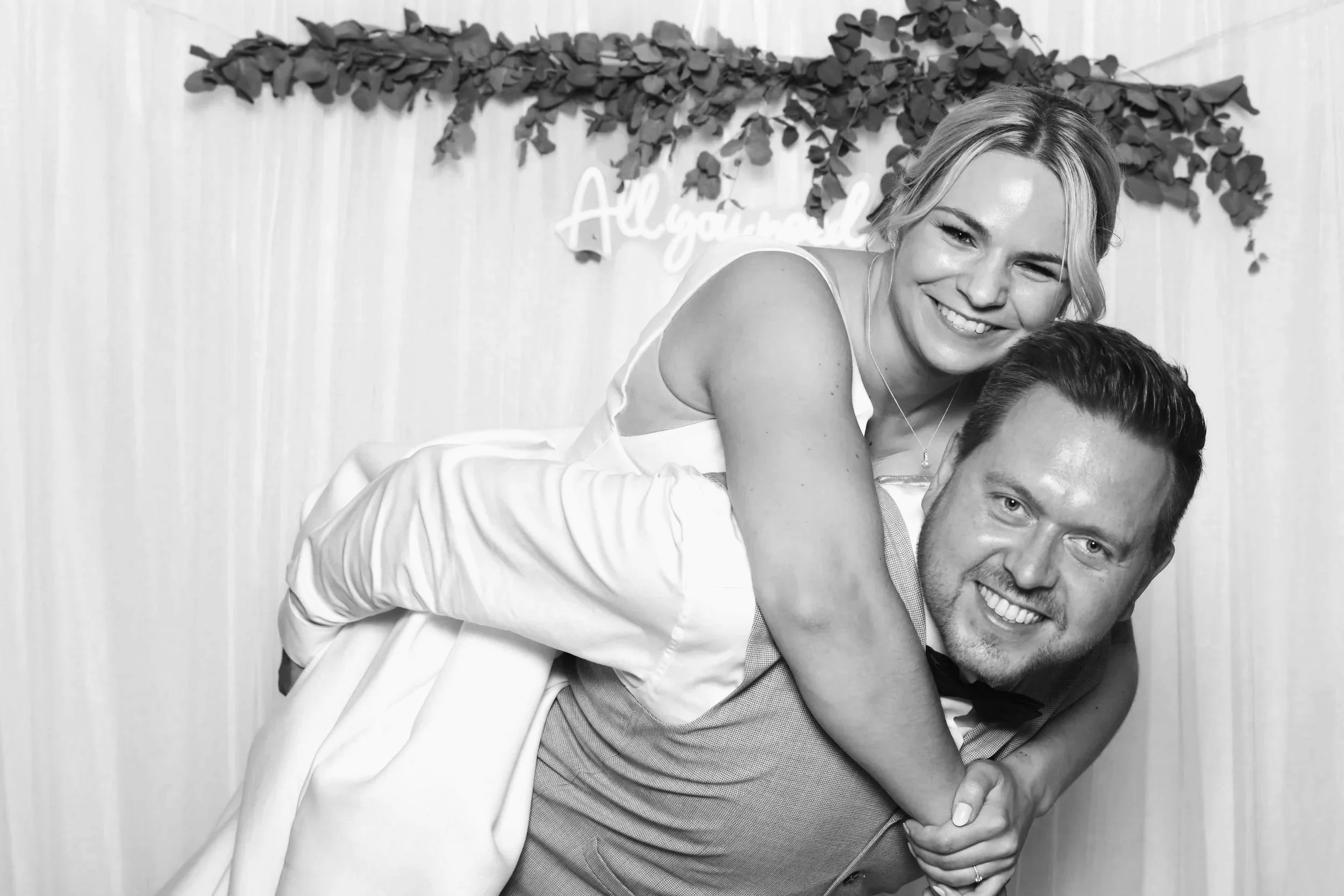 Ein lachendes Paar bei einer Feier, die Frau sitzt auf den Schultern des Mannes, im Hintergrund hängt eine Dekoration mit Blättern und einer Lichtbox mit dem Text 'All you need is love'