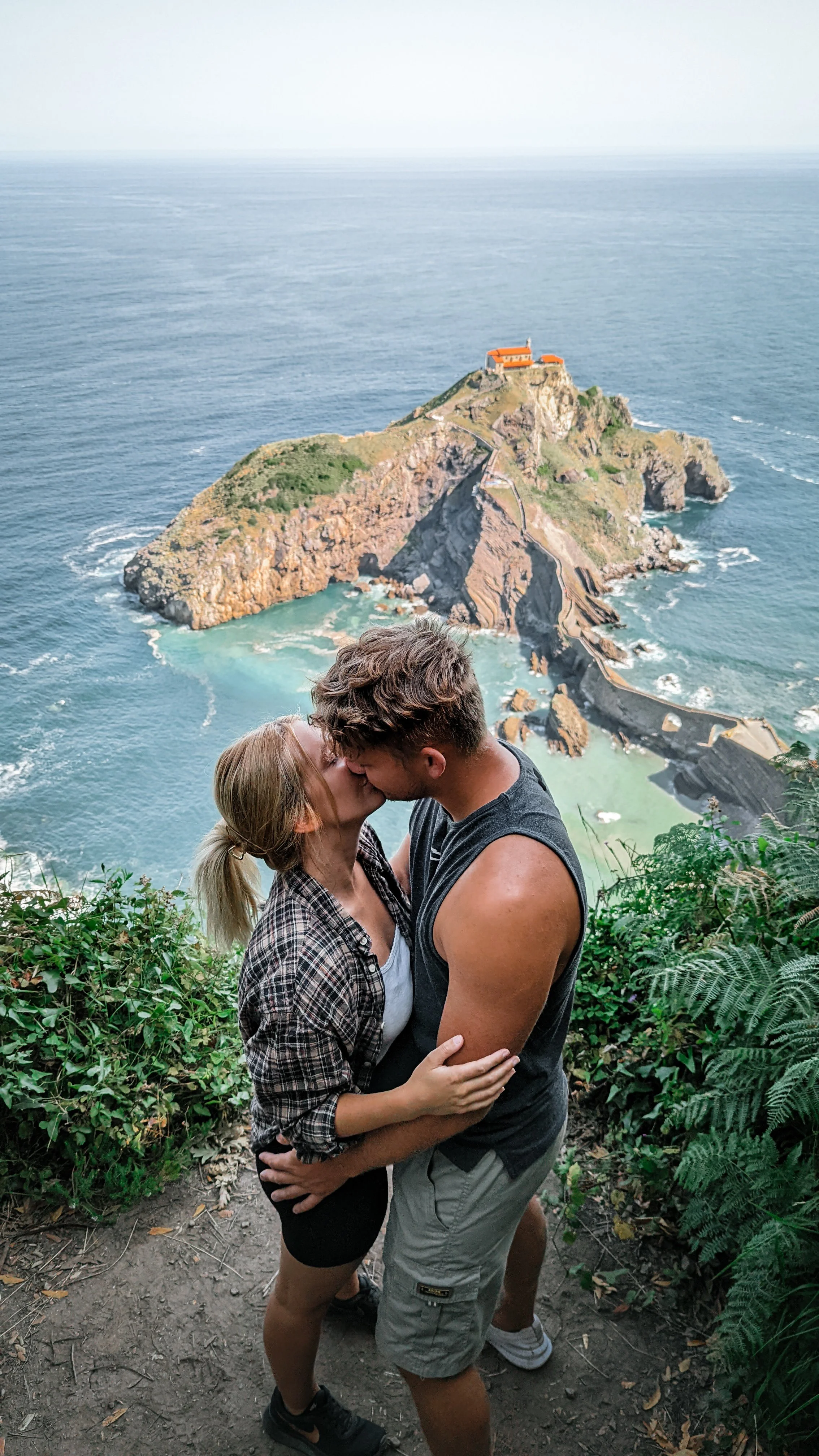 Ein Paar küsst sich verliebt vor einer Klippe mit Blick auf das Meer und eine kleine Insel mit Felsen und Gebäuden.