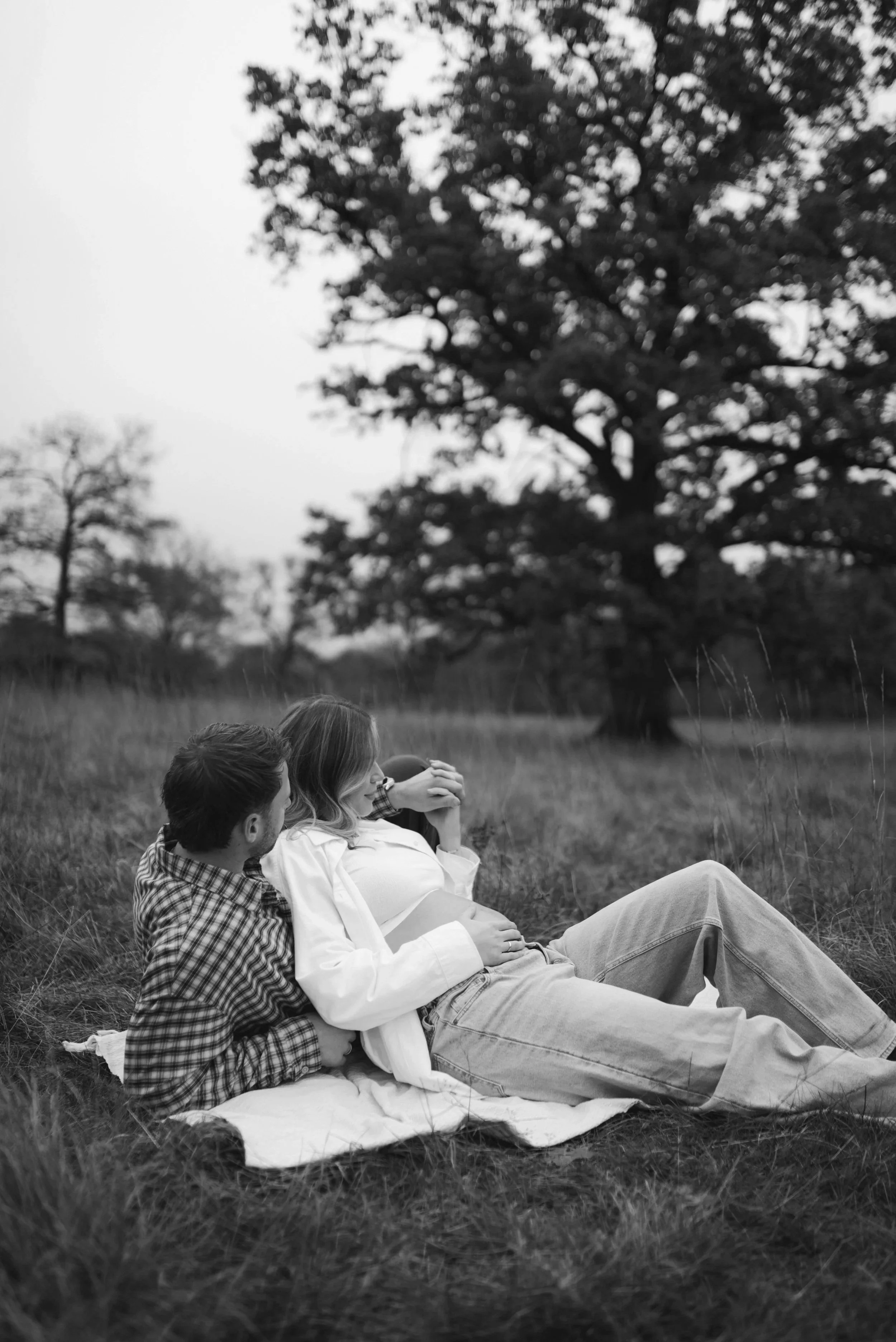 Ein Mann und eine schwangere Frau sitzen gemeinsam auf einer Decke im Freien, mit einem großen Baum im Hintergrund.