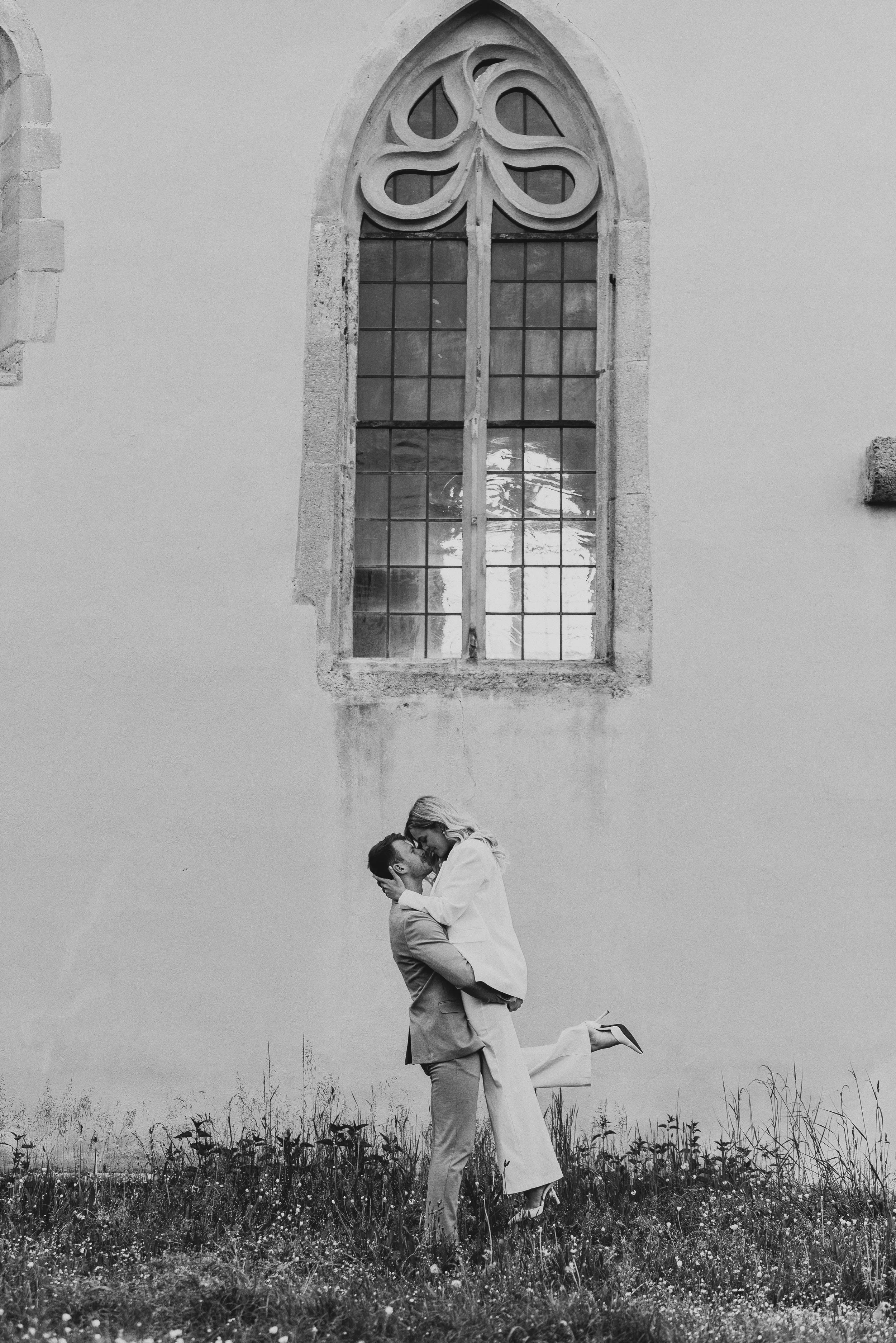 Ein Mann hebt eine Frau in die Luft, während sie sich küssen, vor einer alten Kirchenwand mit einem großen Fenster.