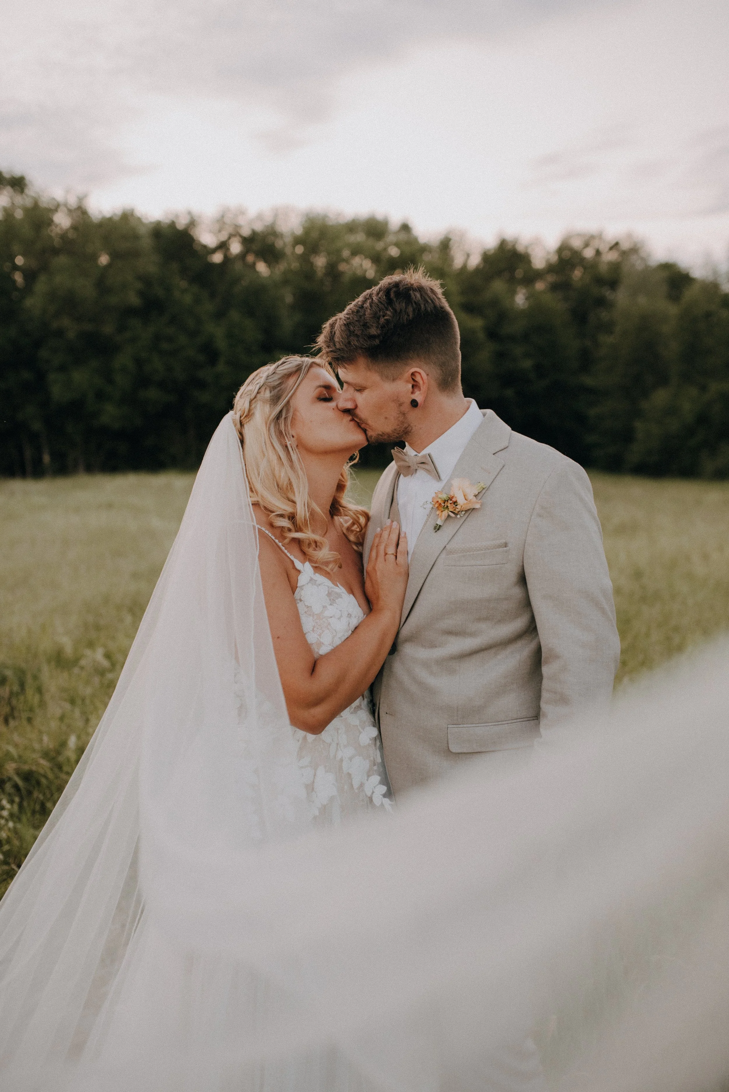 Ein Brautpaar auf dem Feld, das einen Kuss teilt, mit Bäumen im Hintergrund.