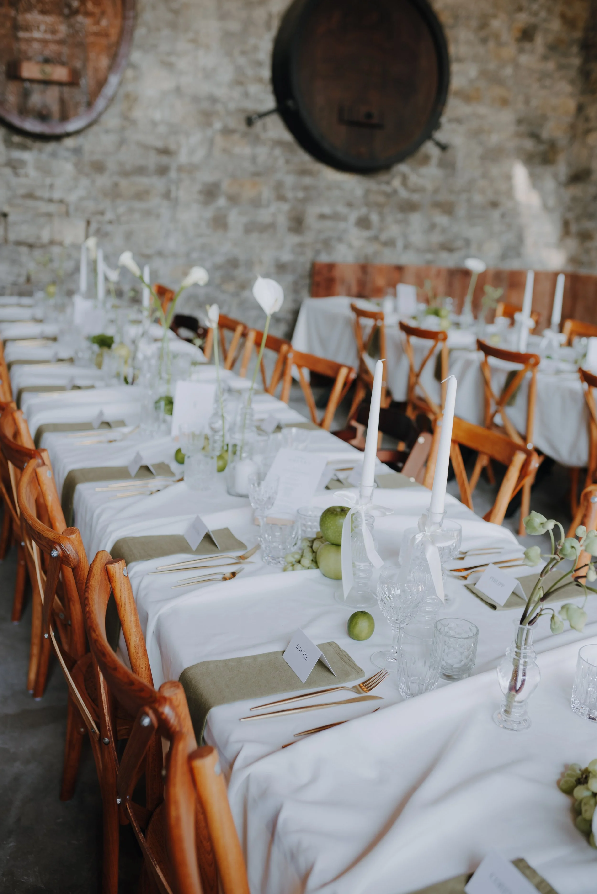 Estrichgedeckter Festsaal mit festlich gedeckten Tischen, weißen Tischtüchern, Kerzen, grünen Äpfeln und Blumenarrangements, umgeben von Holzhockern.