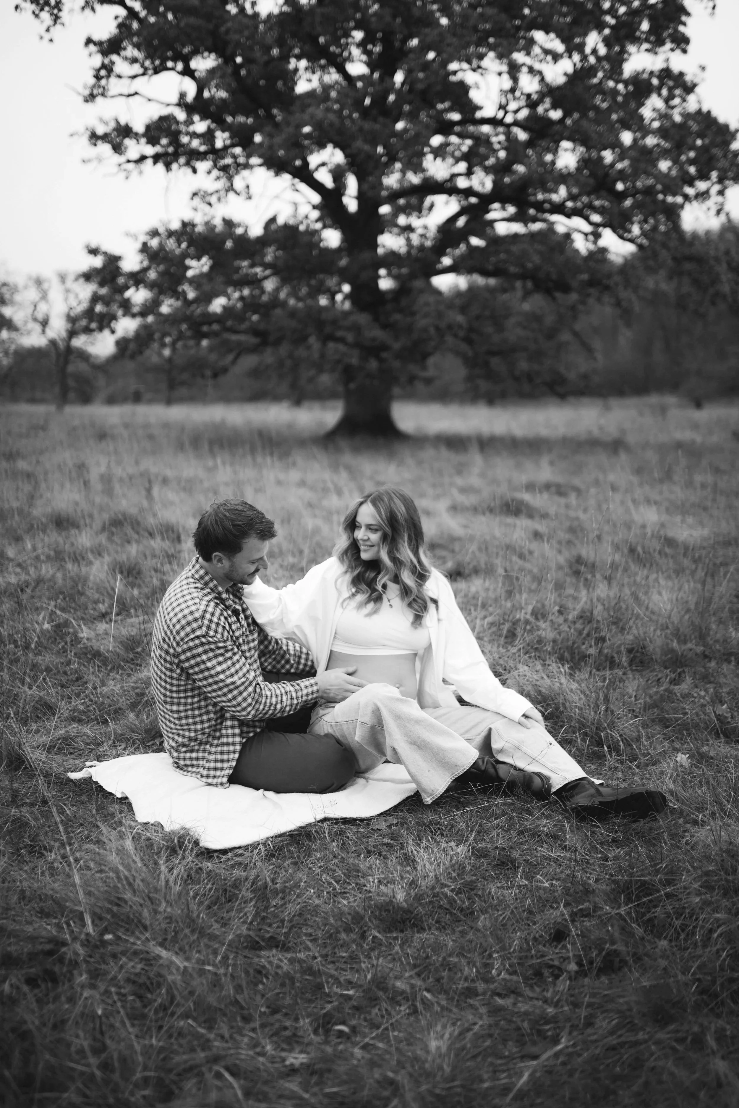 Ein Mann und eine schwangere Frau sitzen auf einer Decke im Gras in einem offenen Feld, umgeben von Bäumen, und lachen gemeinsam.