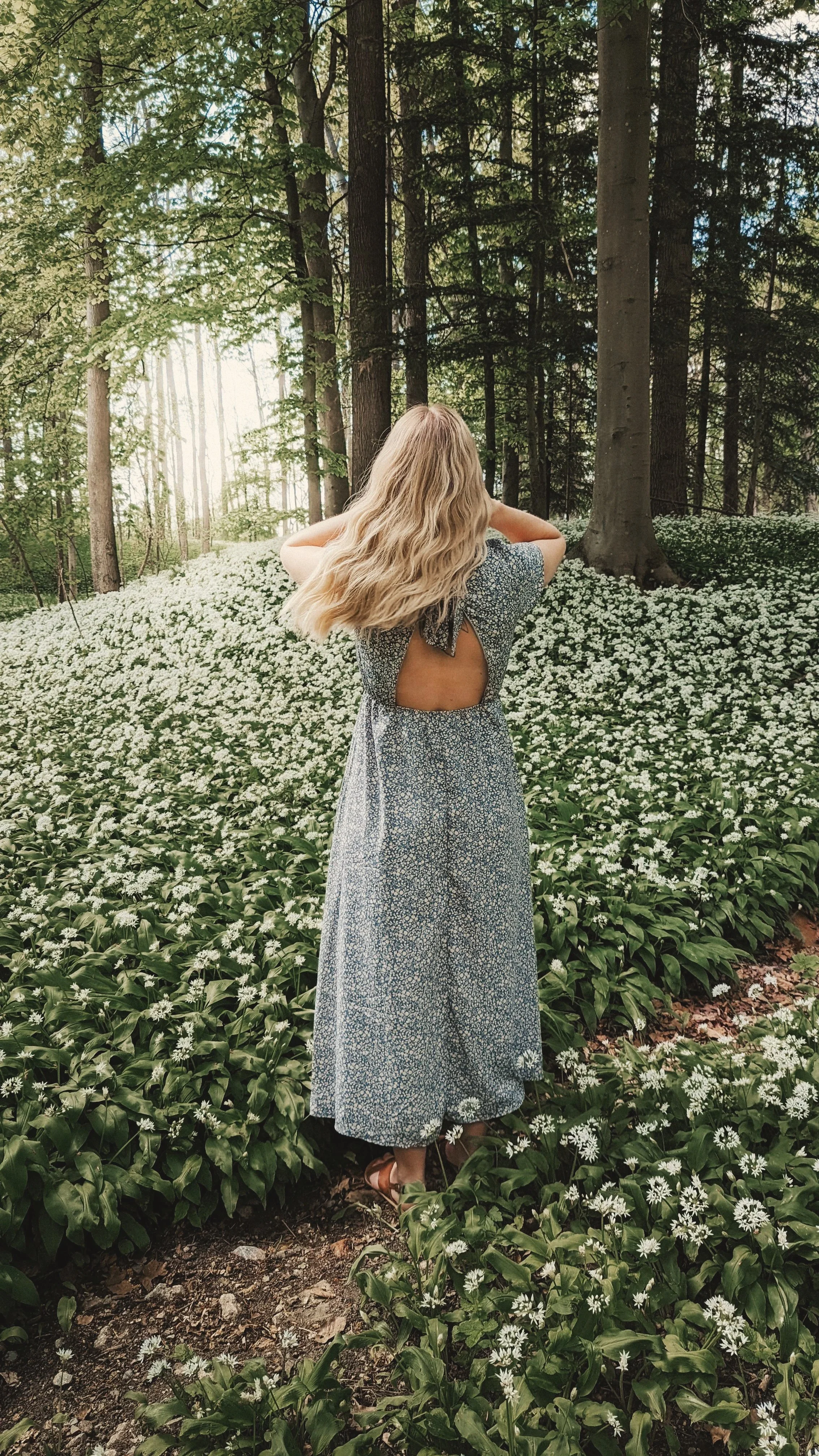 Eine Frau in einem blauen Kleid mit offenem Rücken steht in einem blühenden Wald voller weißer Blumen.