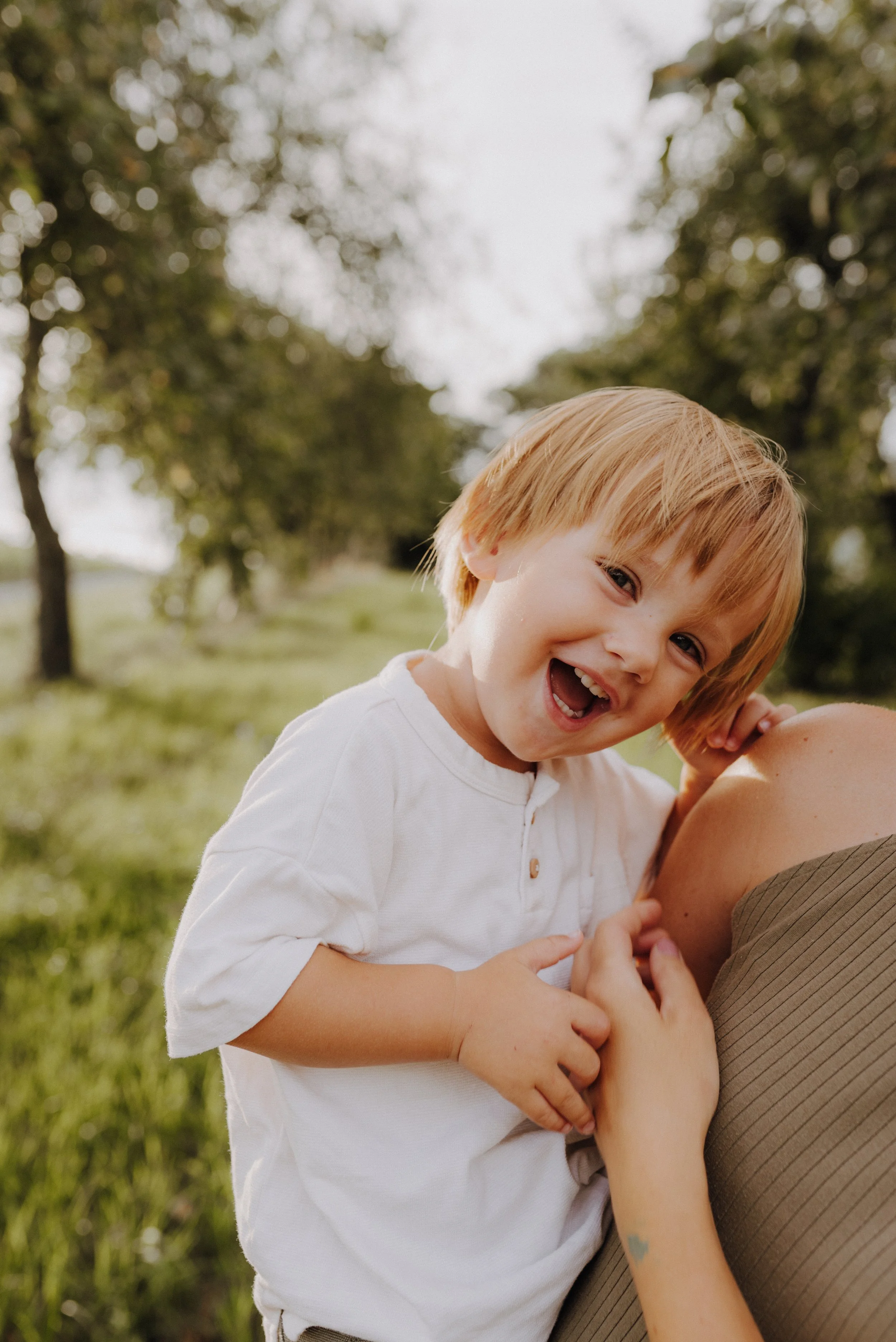 Ein lachendes Kind mit roten Haaren, das auf einer Person auf der Wiese im Freien sitzt.