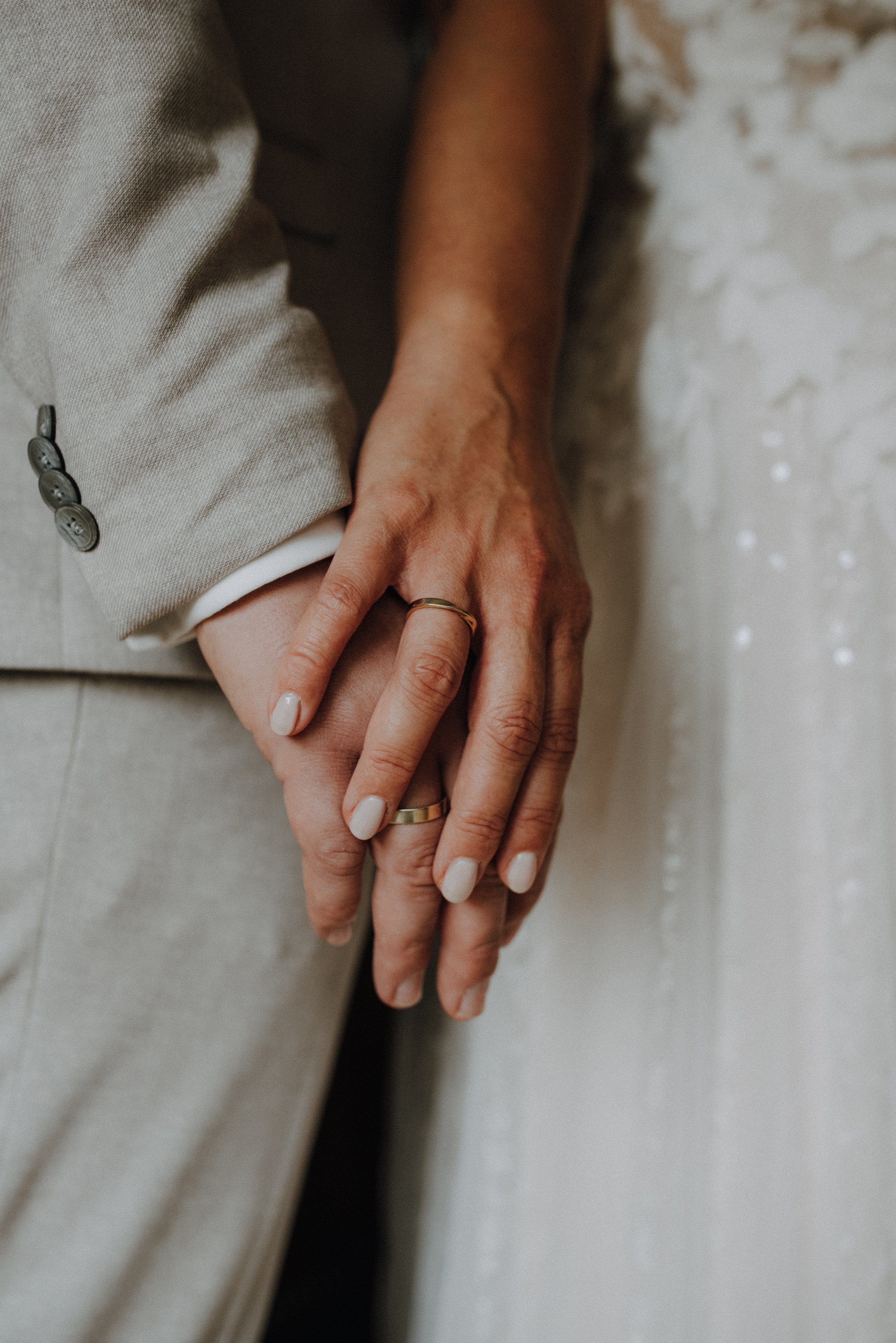 Nahaufnahme von zwei Händen, die sich halten, mit Ringen an den Fingern. Einer trägt einen beige Anzug, der andere ein weißes Kleid.