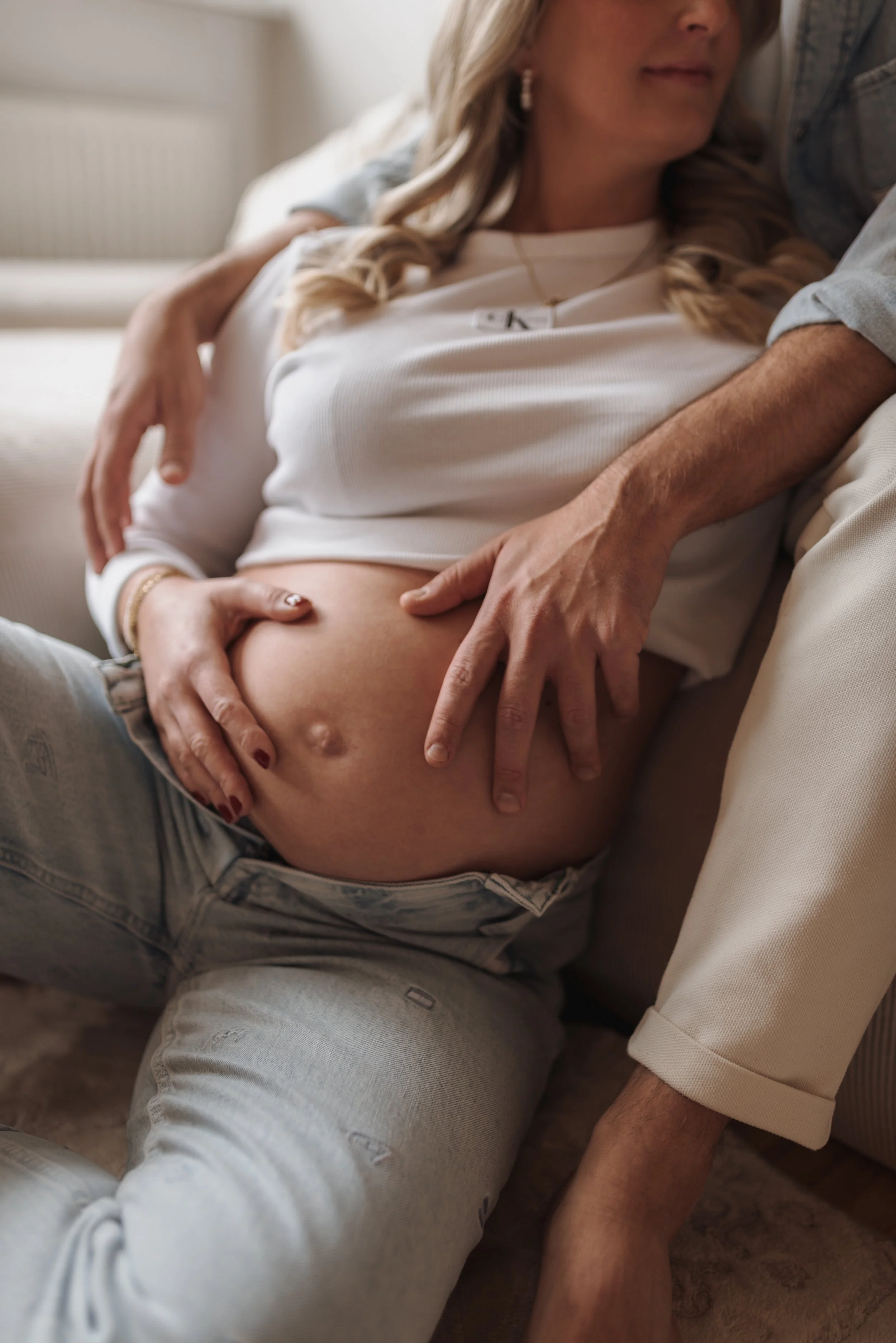 Eine schwangere Frau liegt auf einem Sofa, während jemand ihre Hand auf ihren Bauch legt und sie umarmt.