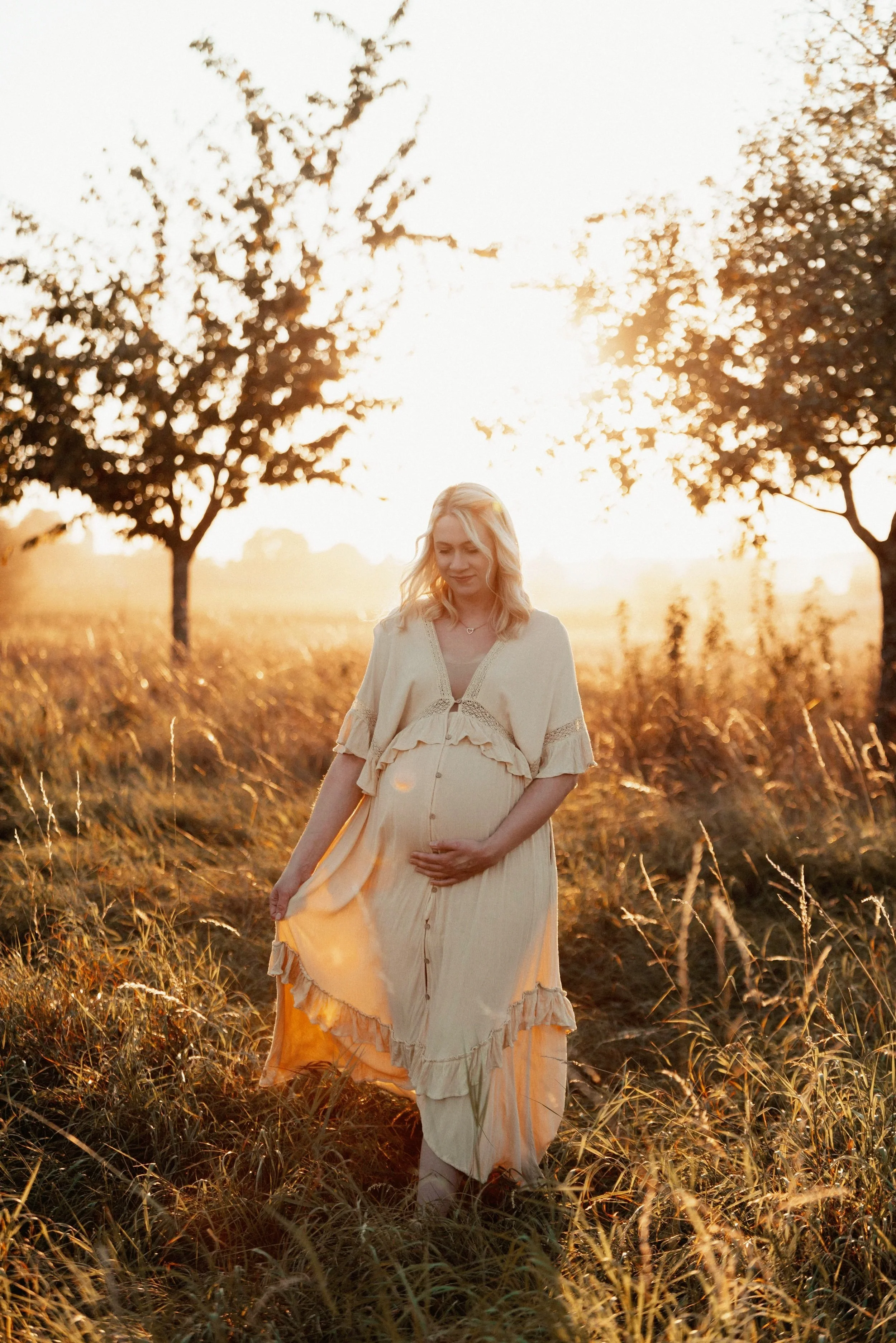 Schwangere Frau in einem weiten, gelben Kleid, die im Sonnenuntergang durch ein Grasdachfeld geht. Im Hintergrund sind Bäume und ein weites, hell erleuchtetes Feld.
