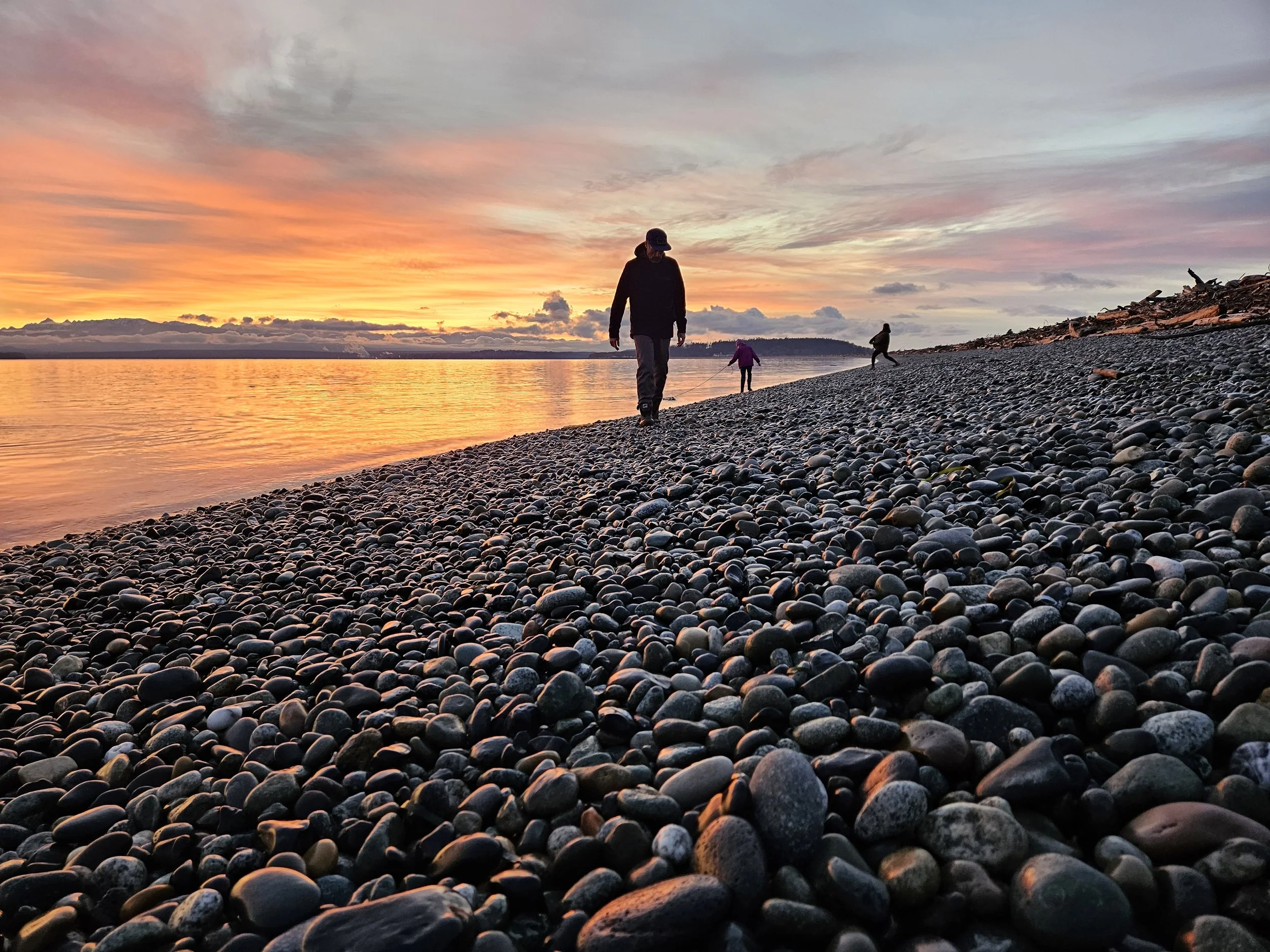 People walking on a pebble beach during sunset with a colorful sky and calm water.
