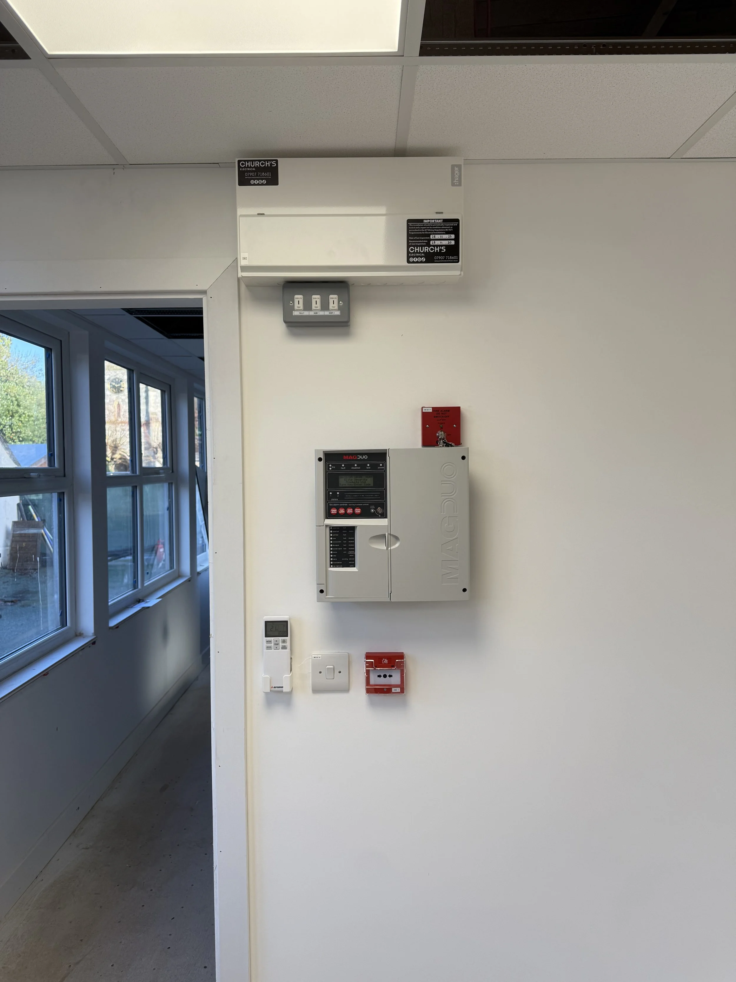 Interior wall with an electrical panel, a fire alarm, a thermostat, and a few switches, next to a hallway with windows.